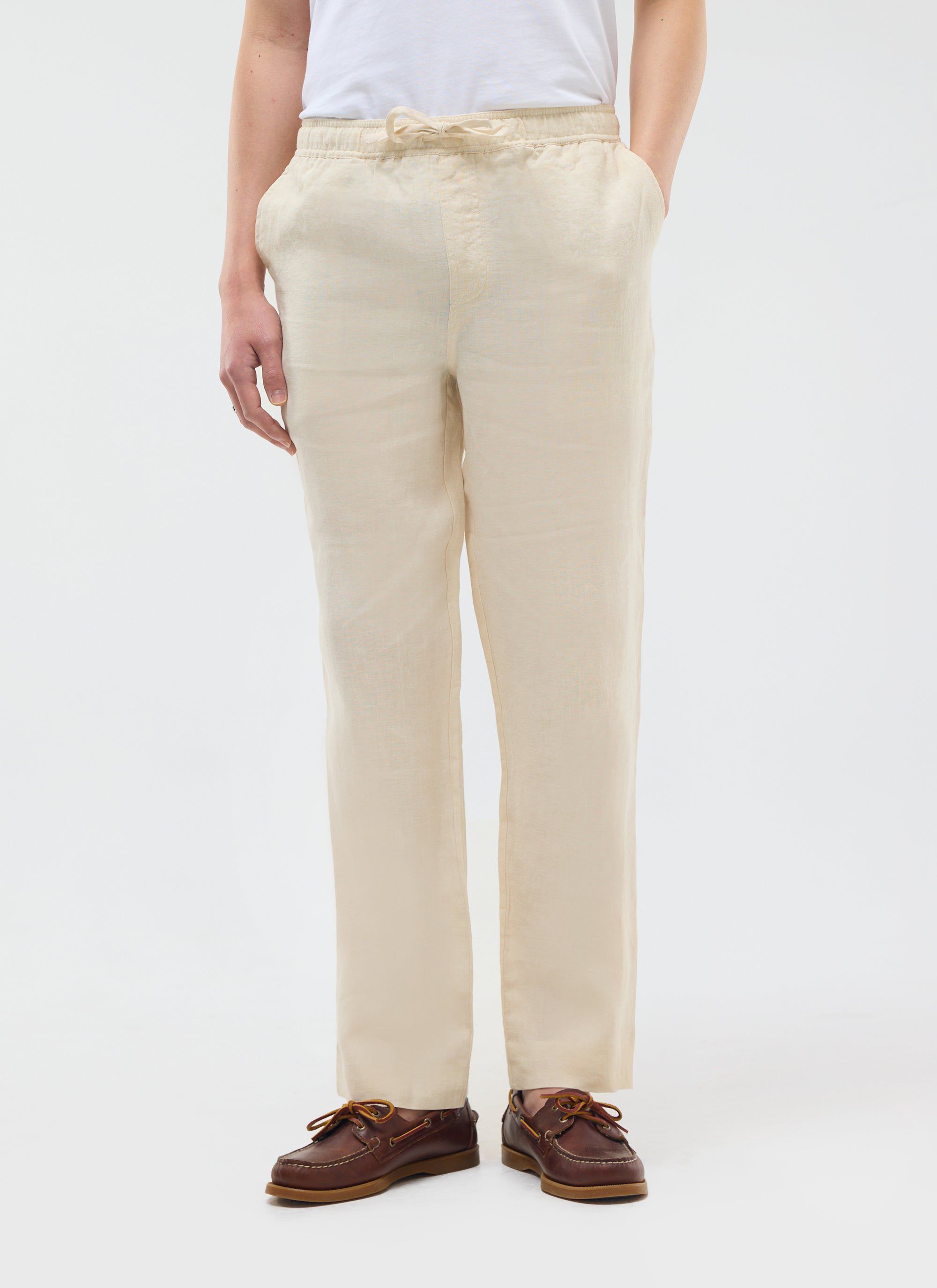 Model wears Percival's cream linen trousers with a white t-shirt and brown boat shoes against a white studio background.