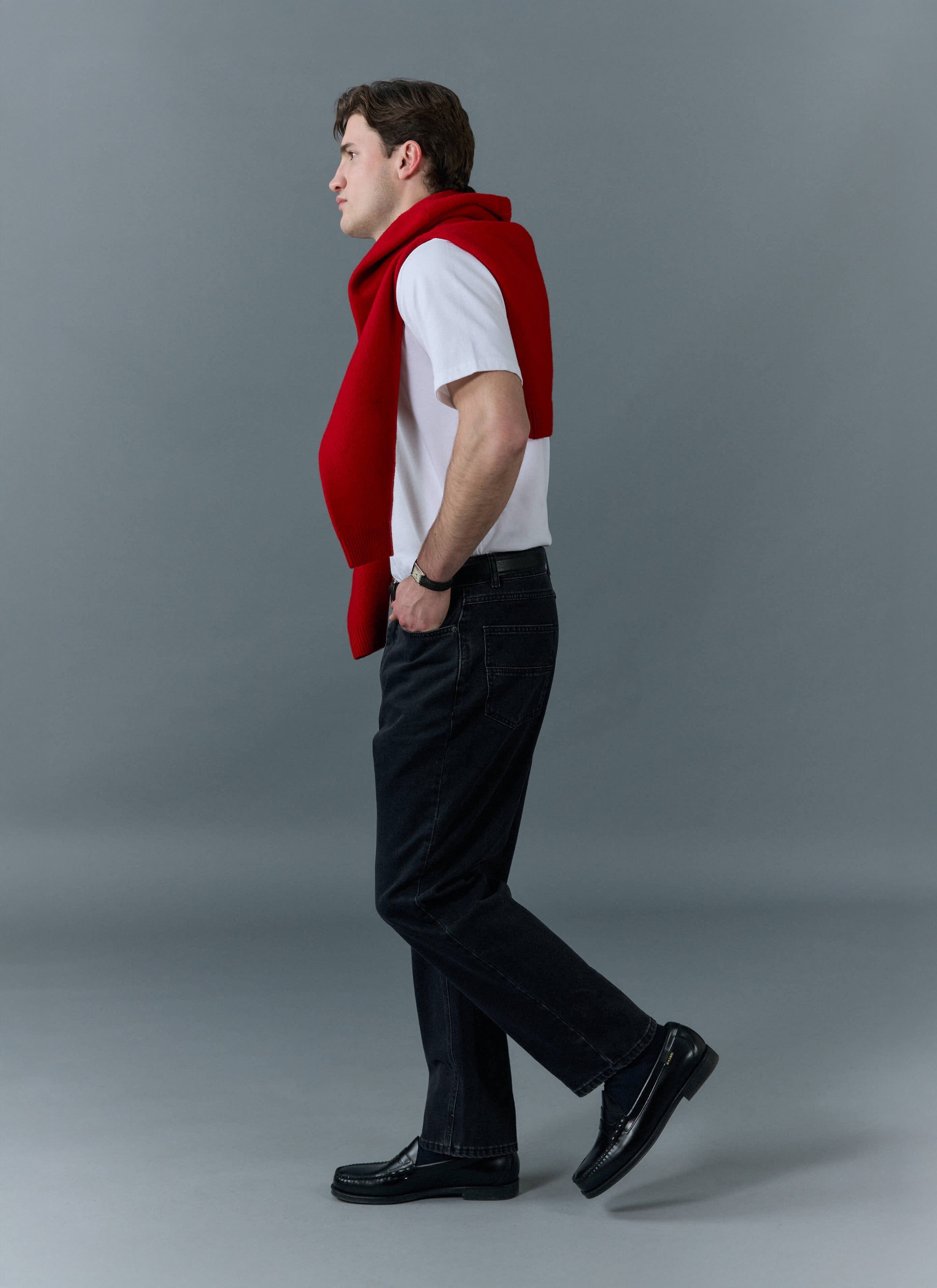 Side profile of a model wearing Percival black straight leg denim jeans, a white t-shirt and a red jumper over his shoulders.