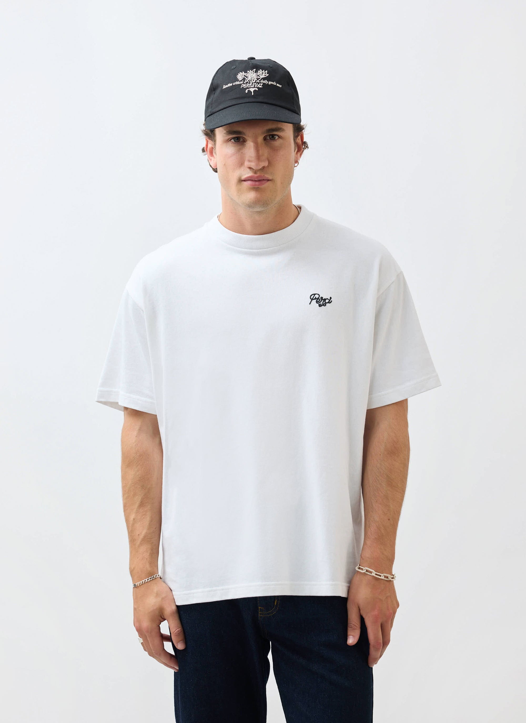 A model wears the Percival Du Temps Oversized T Shirt in white, styled with dark denim and a black cap in a studio.