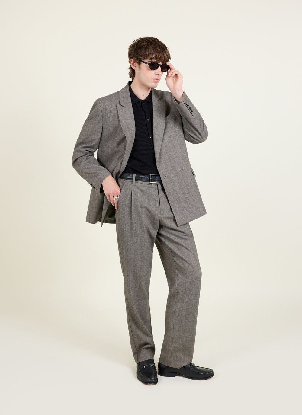 A model wears the Percival grey herringbone tailored suit, styled with a black polo shirt, loafers, and sunglasses.