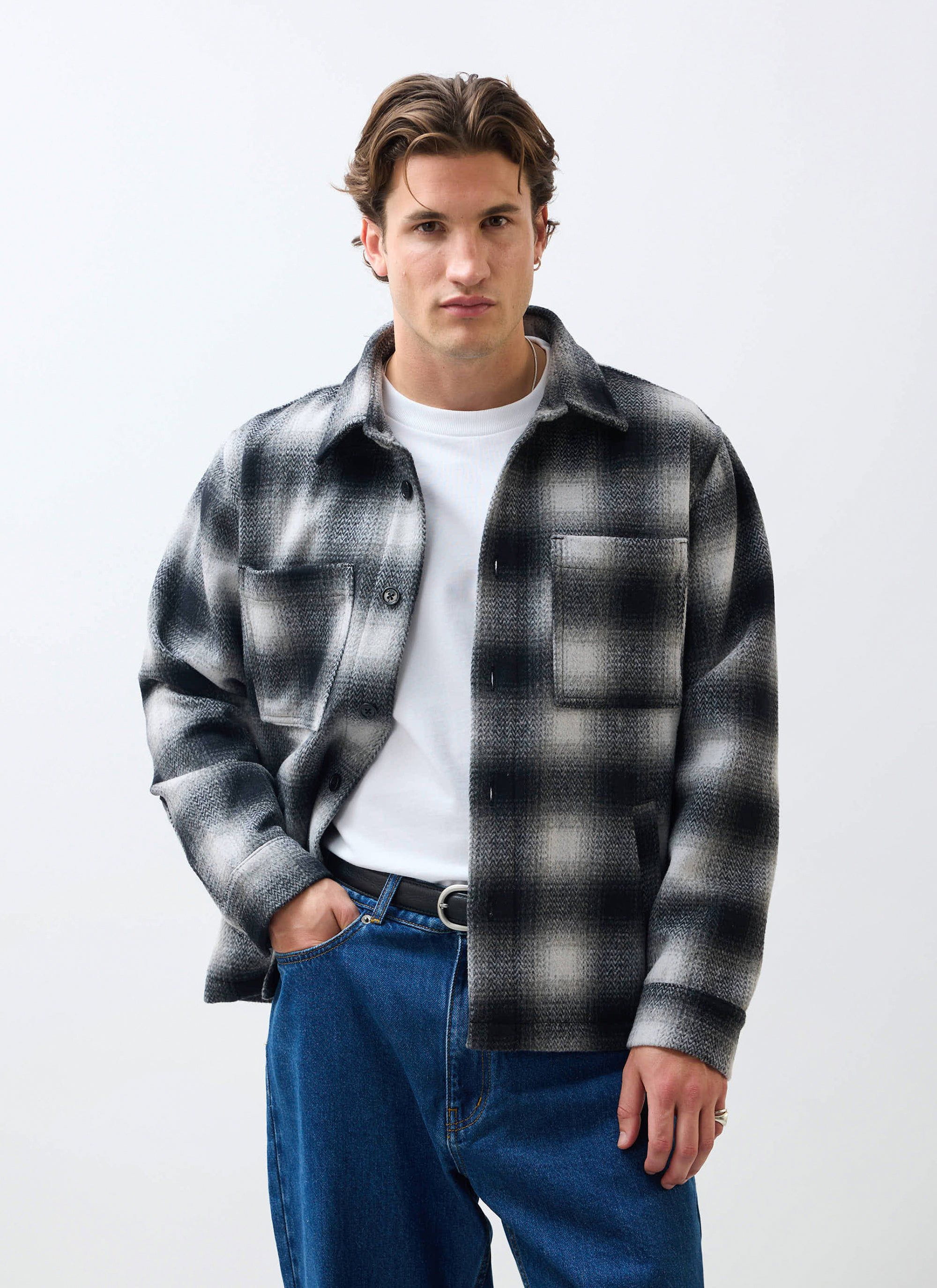 A man models the Percival Check Overshirt in grey, a wool-blend shacket styled over a white t-shirt and dark jeans.