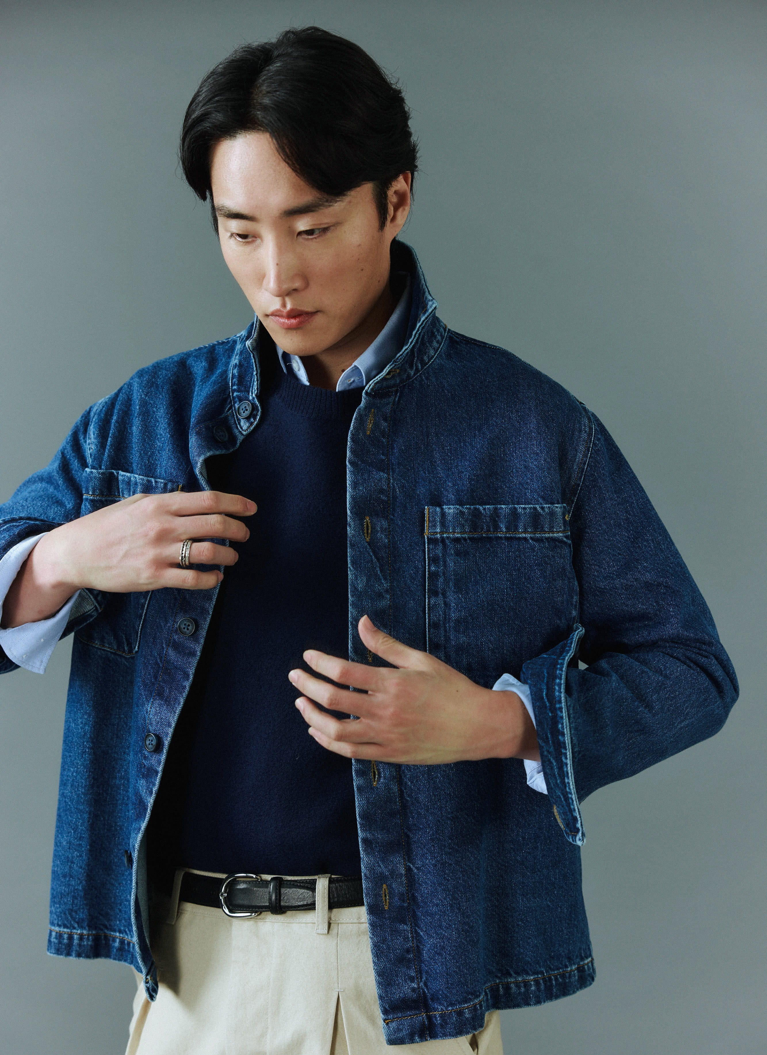 A man models the Percival Felted Regular Jumper in navy merino wool, layered under a denim jacket against a grey wall.