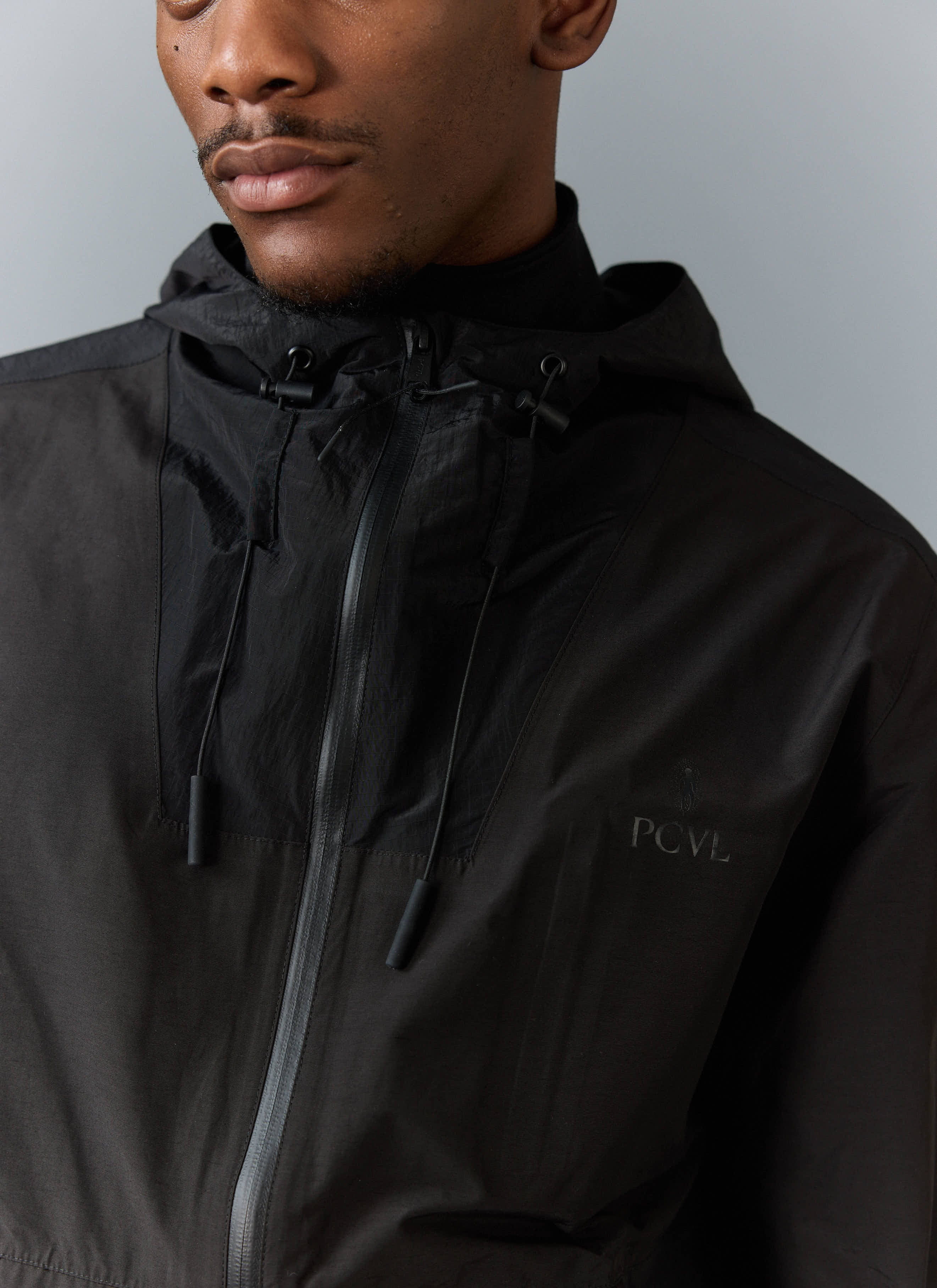A close-up of a model wearing the black PCVL Zip Through Water Resistant Jacket from Percival, focusing on the tonal logo.