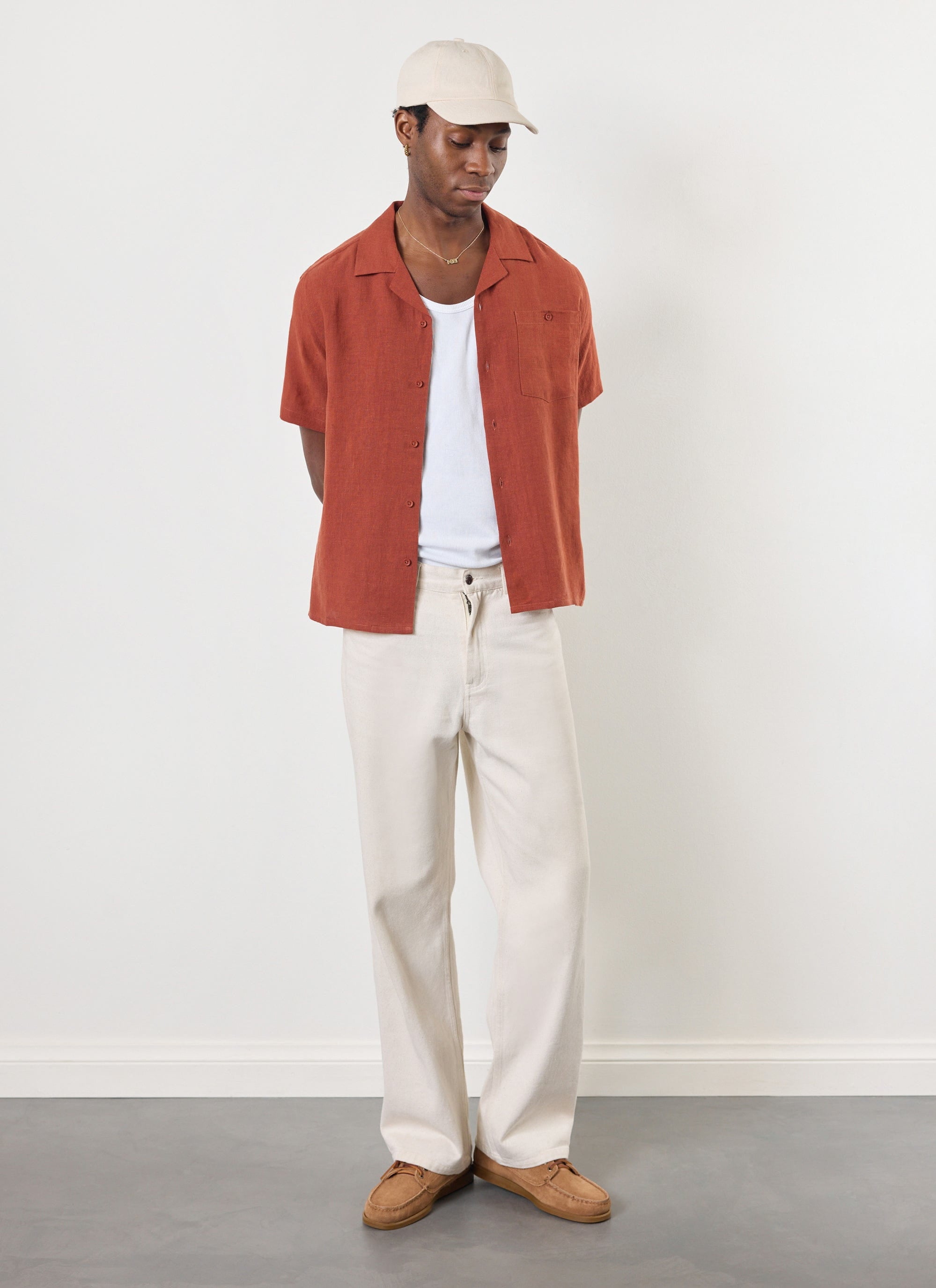 Model wears the Percival rust linen short sleeve Cuban shirt, styled open with a white vest, off-white trousers and a cap.
