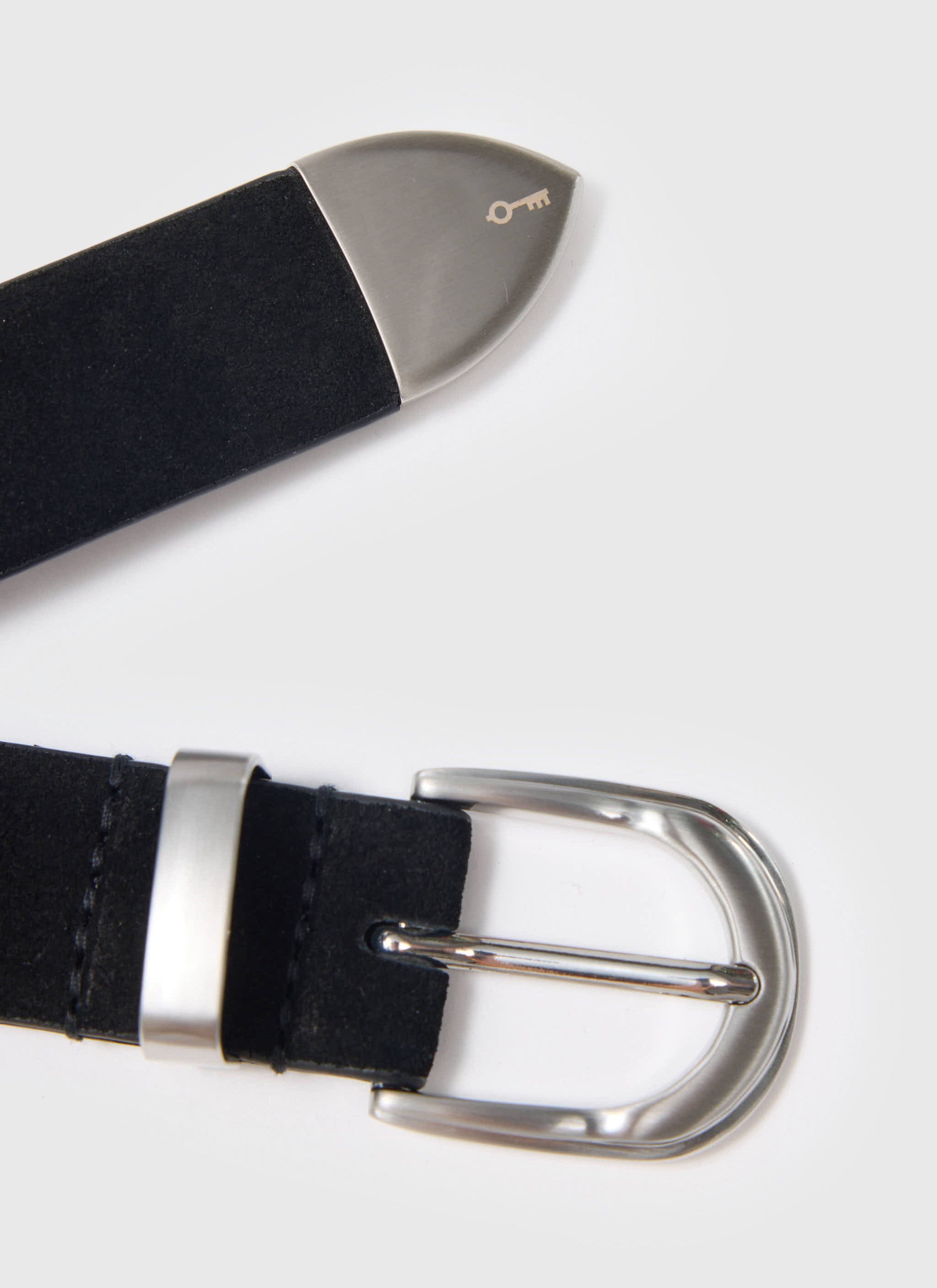 A close-up of the black Suede Belt from Percival, showing the silver buckle and the engraved key logo on the silver tip.