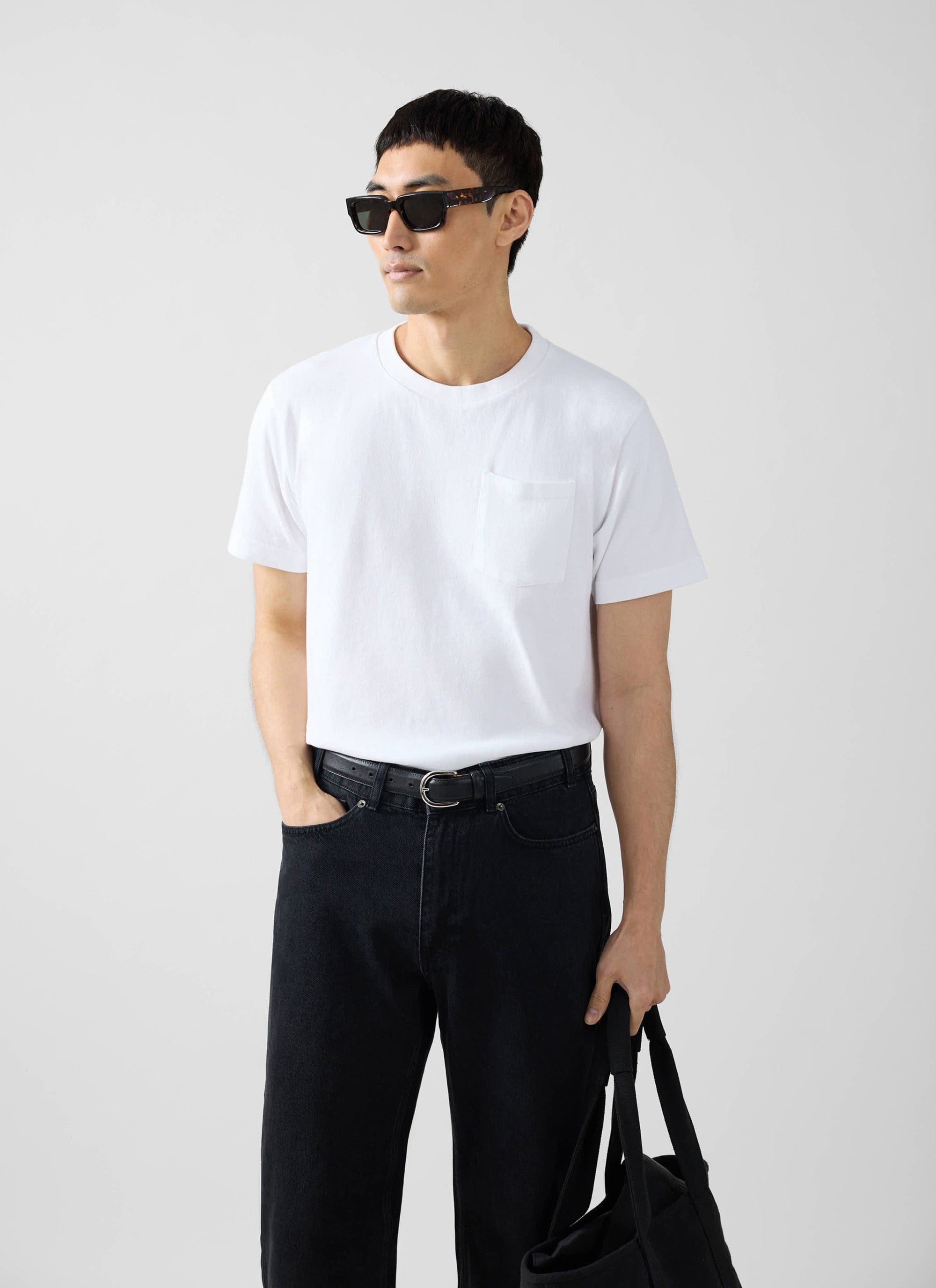 A man models the Percival Short Sleeve Pocket T-Shirt in white with black jeans, sunglasses, and a tote bag.