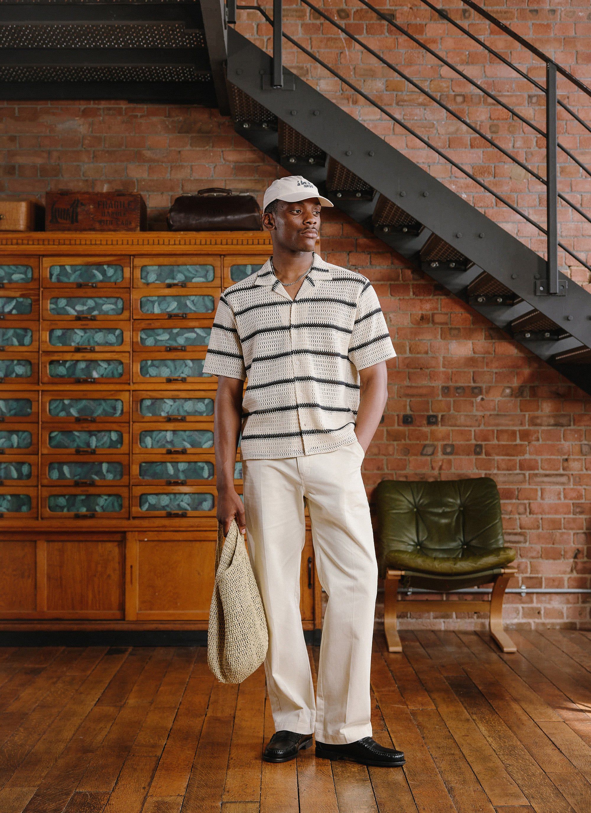 A model wears the Percival Crochet Groundsman Cuban Shirt in ecru with black stripes, with matching trousers and a cap.