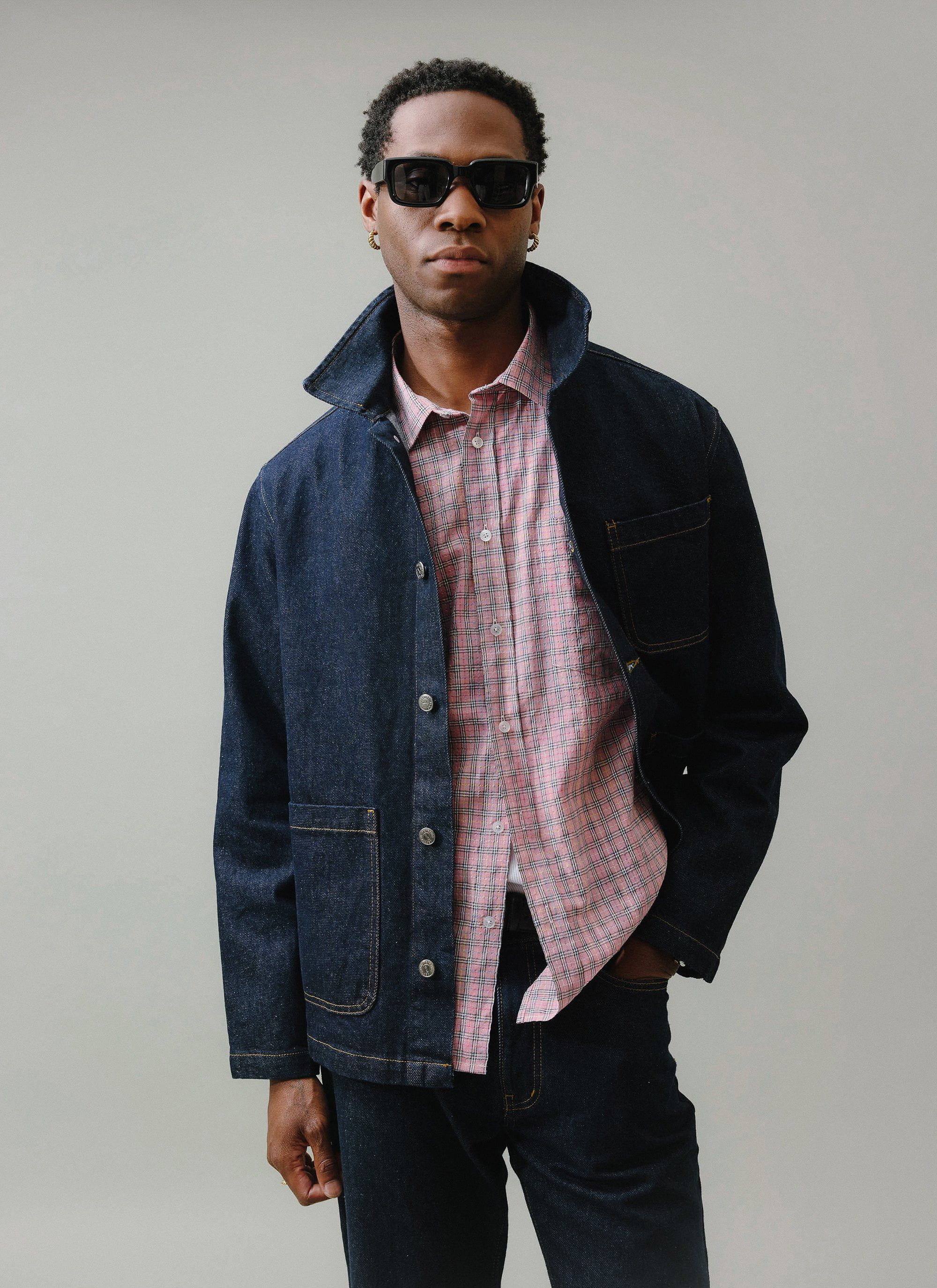 A model wears the Percival Gove Chore Jacket in dark indigo, styled open over a pink checked shirt with matching denim jeans.