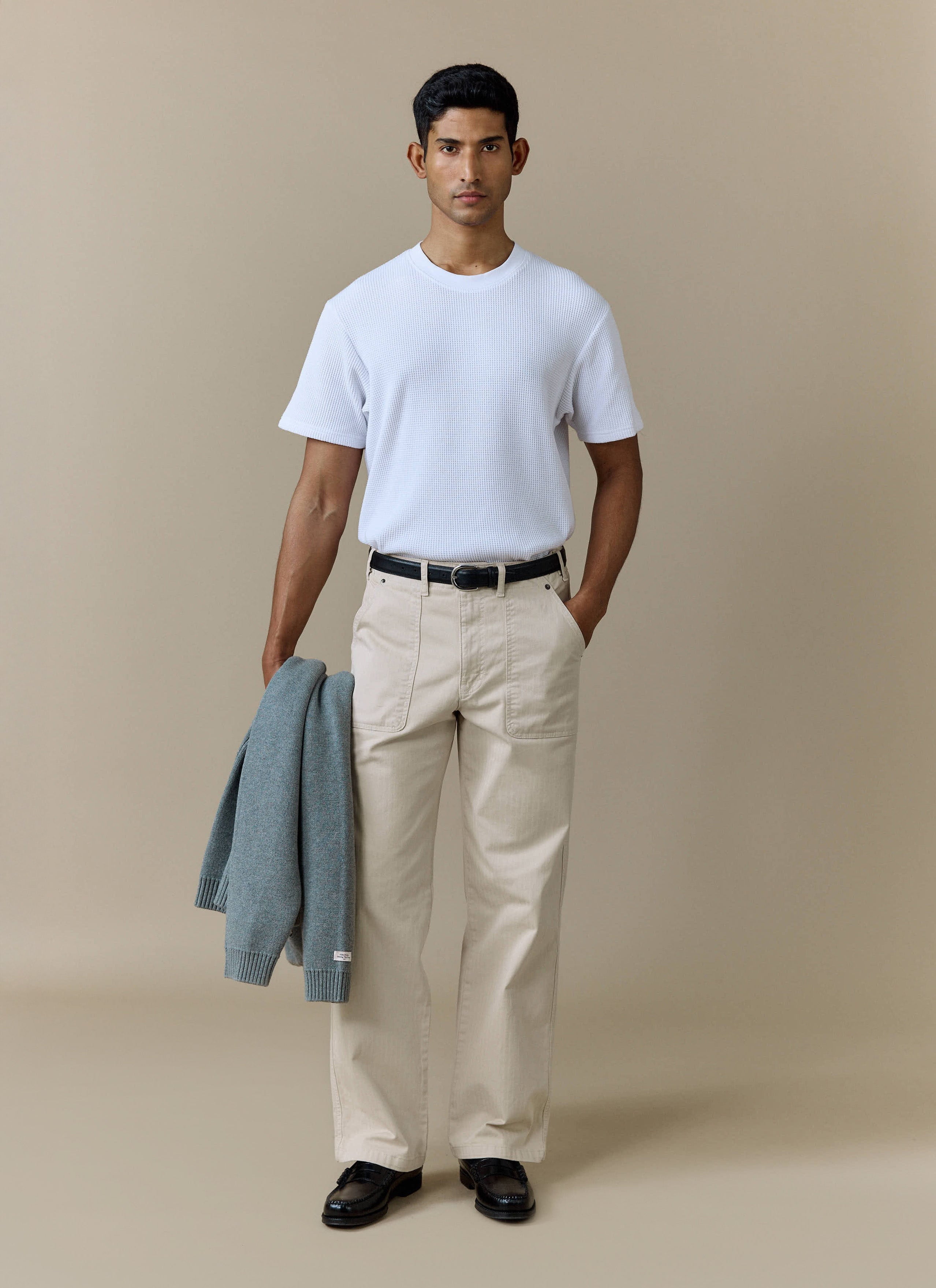 A model wearing the Percival Lungo Herringbone Straight Trousers in cream cotton, holding a grey knit cardigan.