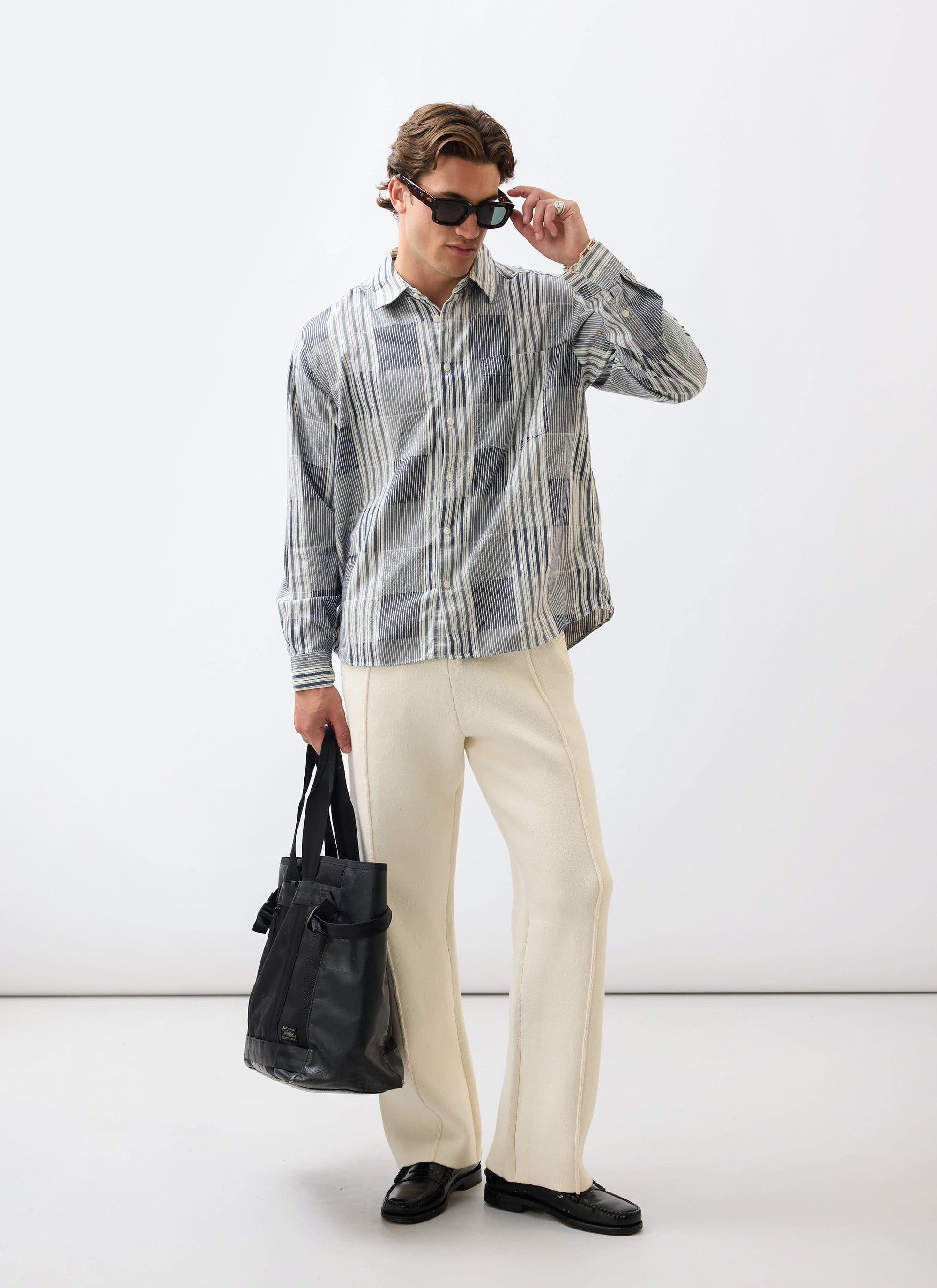 A model wears the Percival Epigraph Patchwork Pearce shirt in blue with cream trousers, holding a black bag and sunglasses.