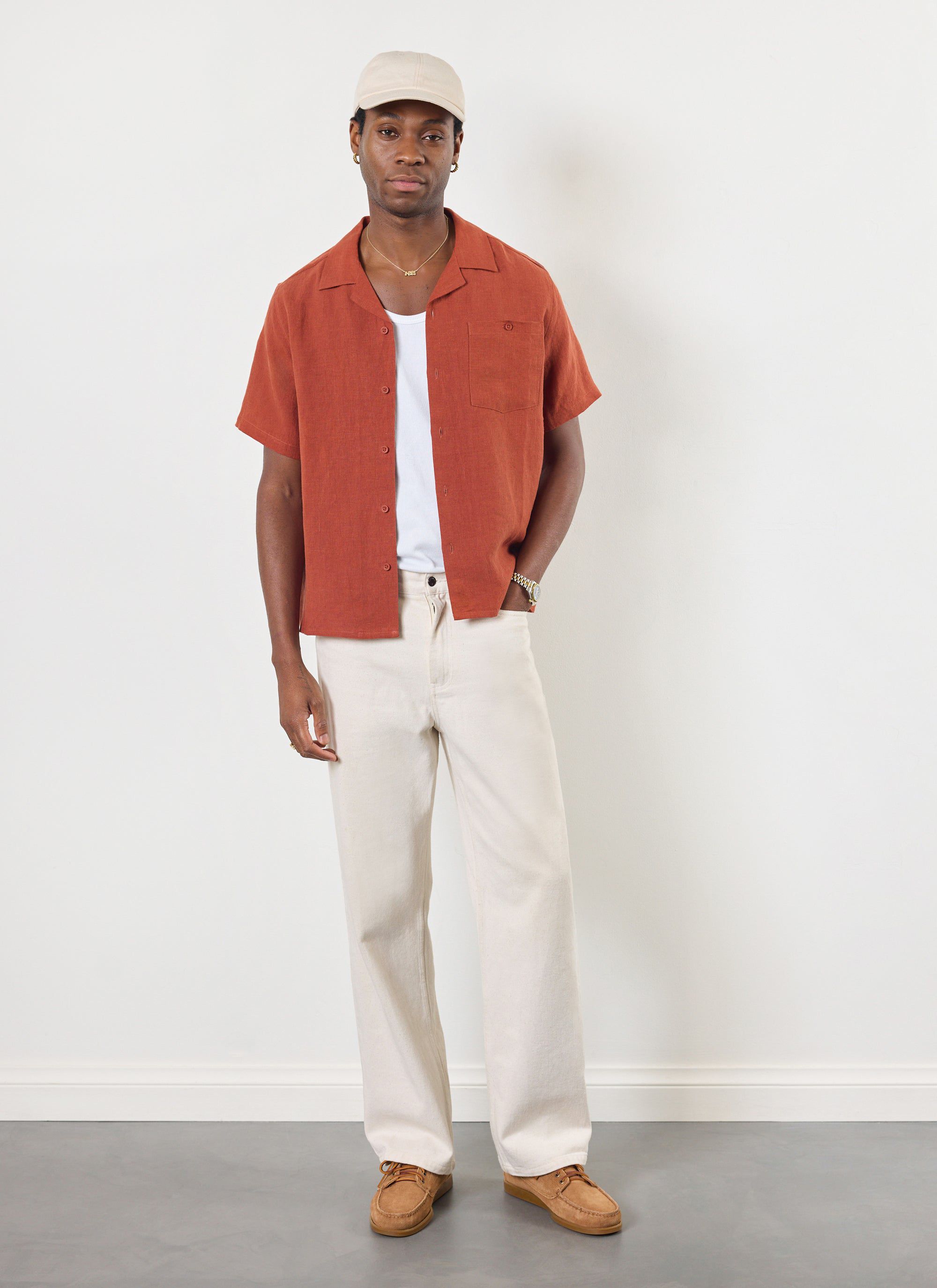 A model wears the Percival rust linen short sleeve Cuban shirt open over a white tee with off-white trousers and a cap.