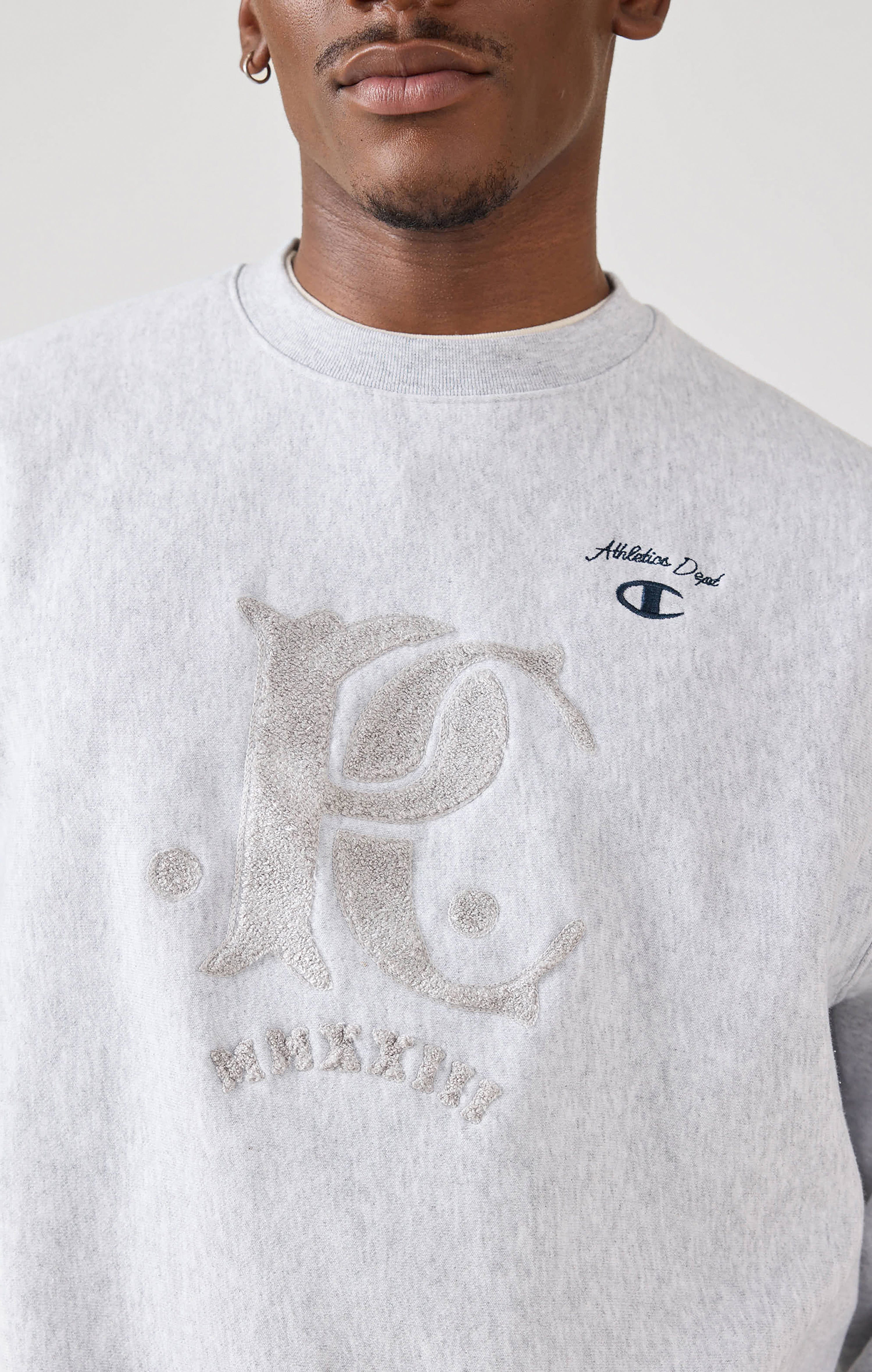 A close-up of a model wearing the grey Percival x Champion Chenille Patch sweatshirt, showing the textured logo detailing.