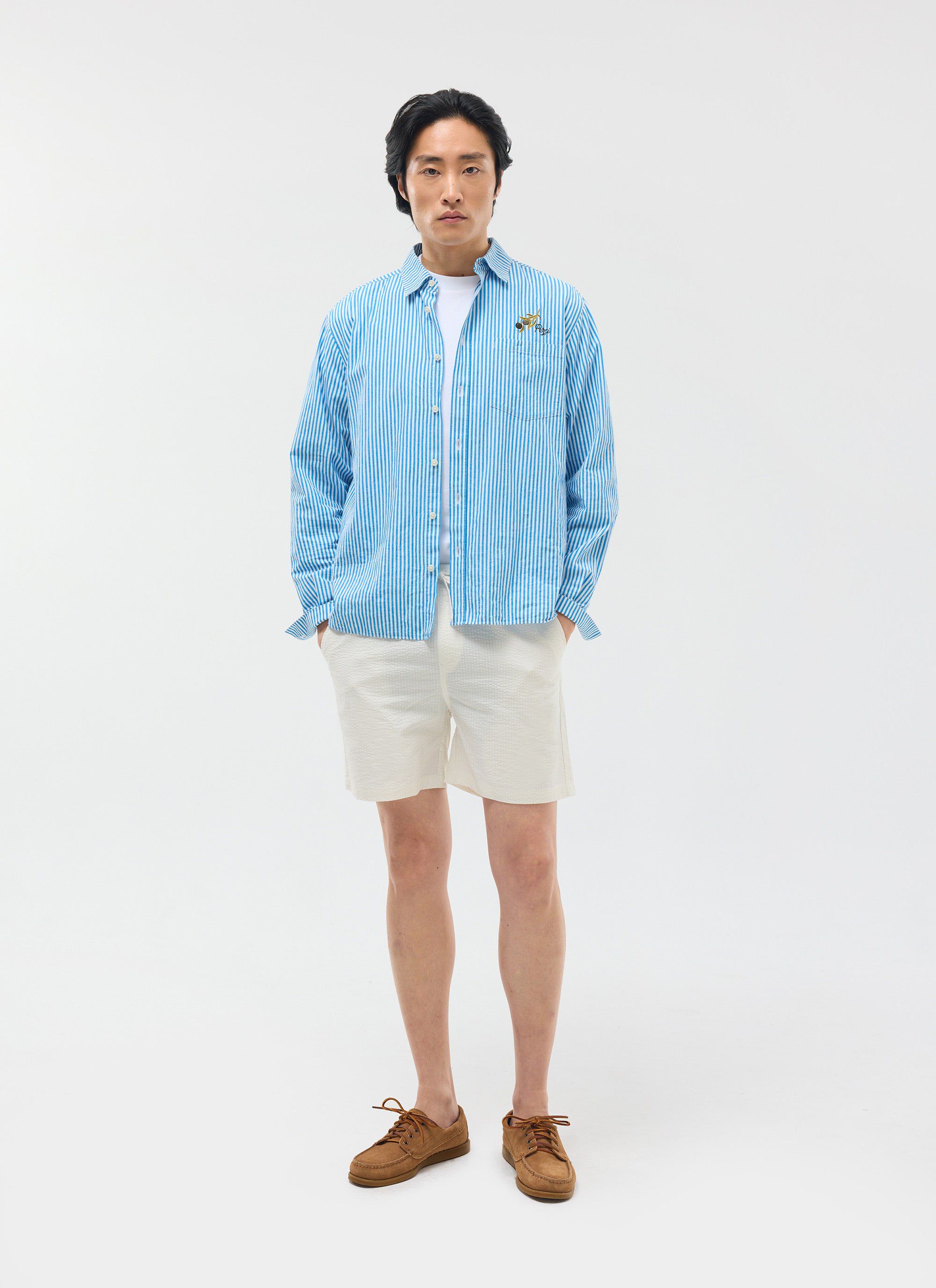 A model wears the Percival Classic Shorts in cream seersucker, styled with a blue striped shirt and brown shoes.