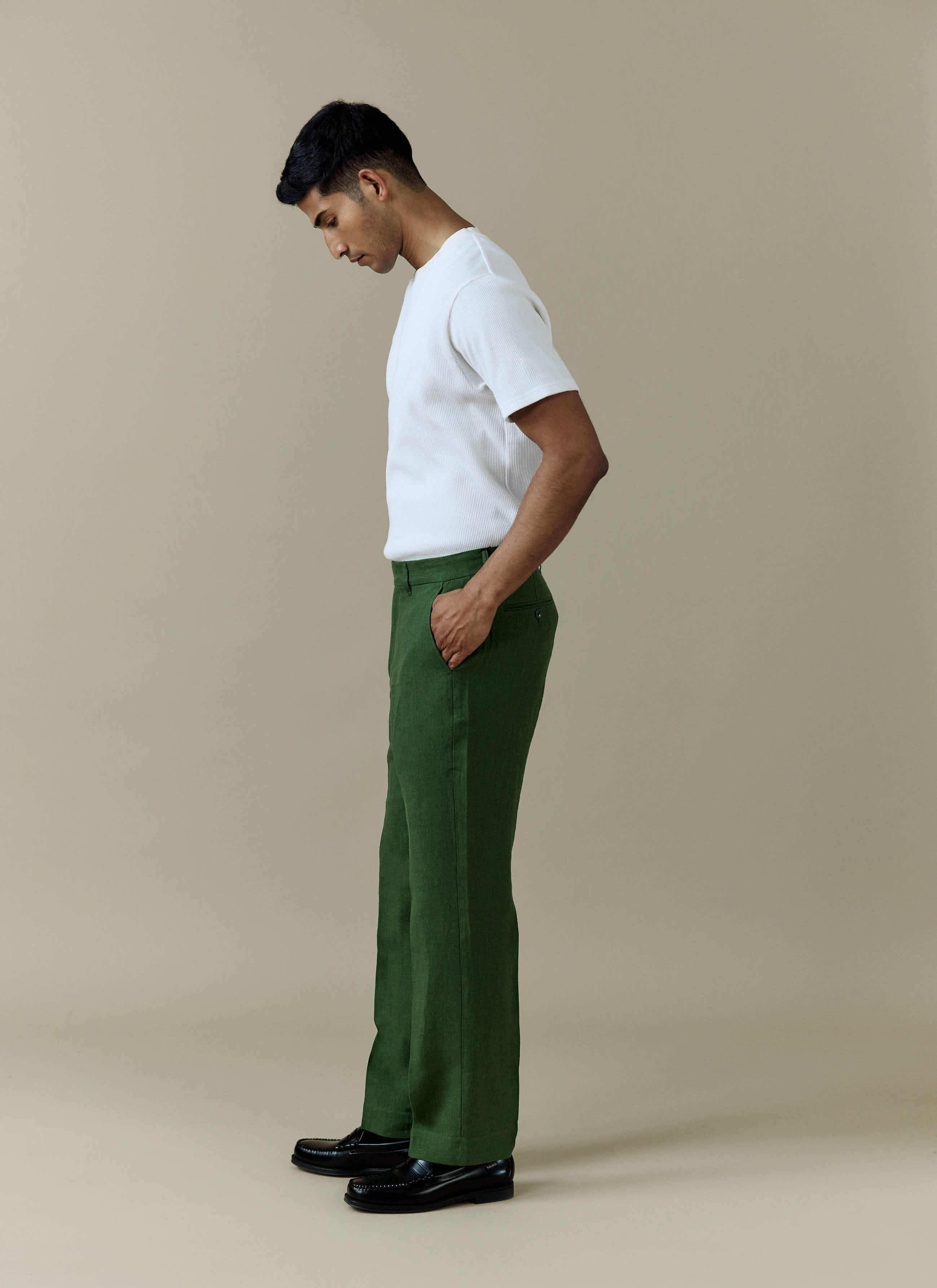 Side view of a model wearing Percival Tailored Linen Trousers in forest green with a white t-shirt and black loafers.