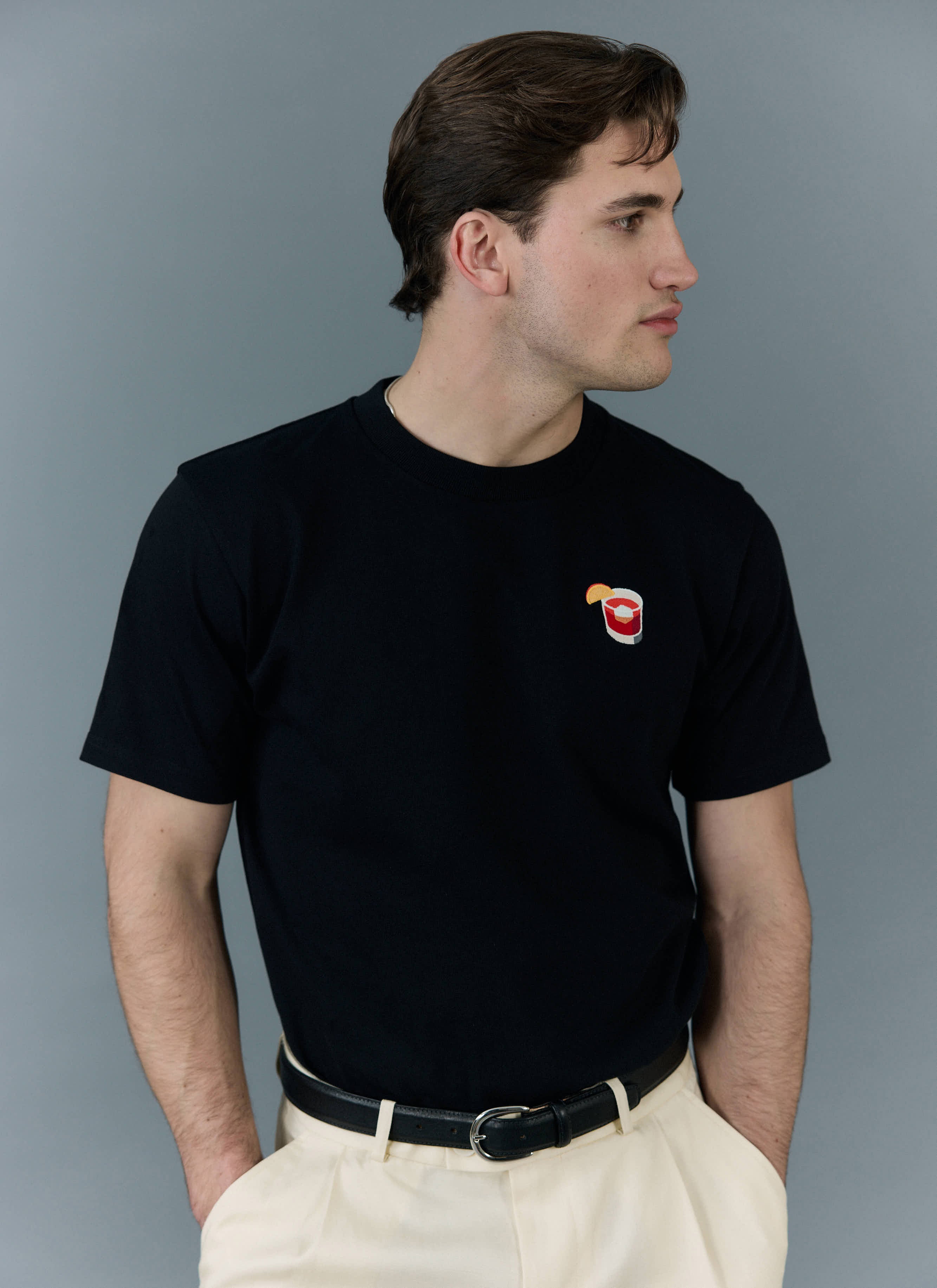 A model wears the black Percival Negroni t-shirt with intricate cocktail embroidery, styled with cream trousers.
