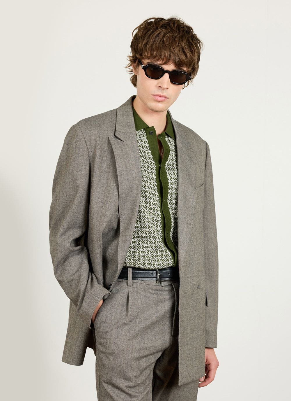 A model wears the Percival grey herringbone double breasted blazer with matching trousers, a green knit shirt and sunglasses.