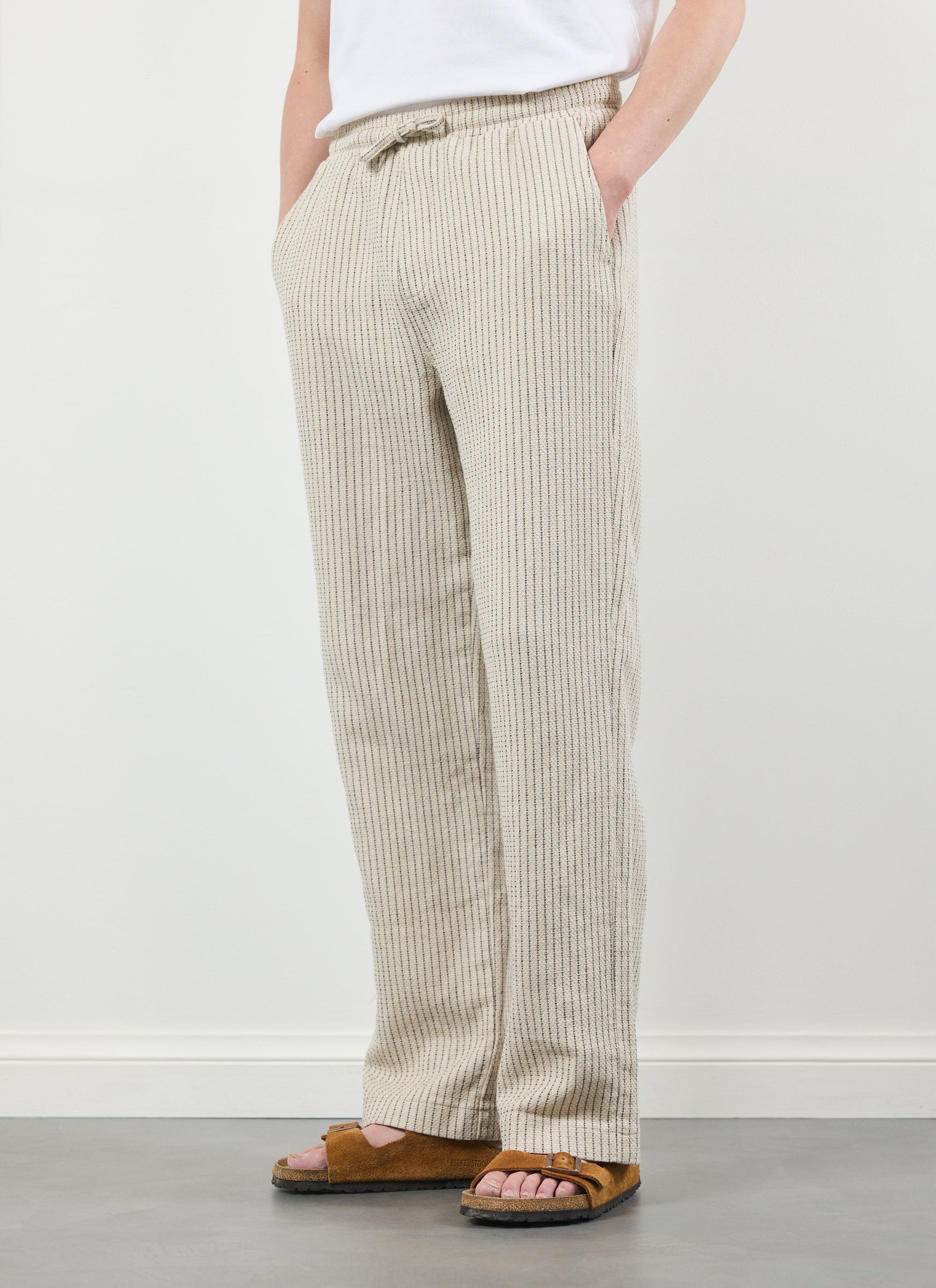 A model wears the ecru striped Sorrento Linen Trousers from Percival, standing with hands in pockets in a studio.