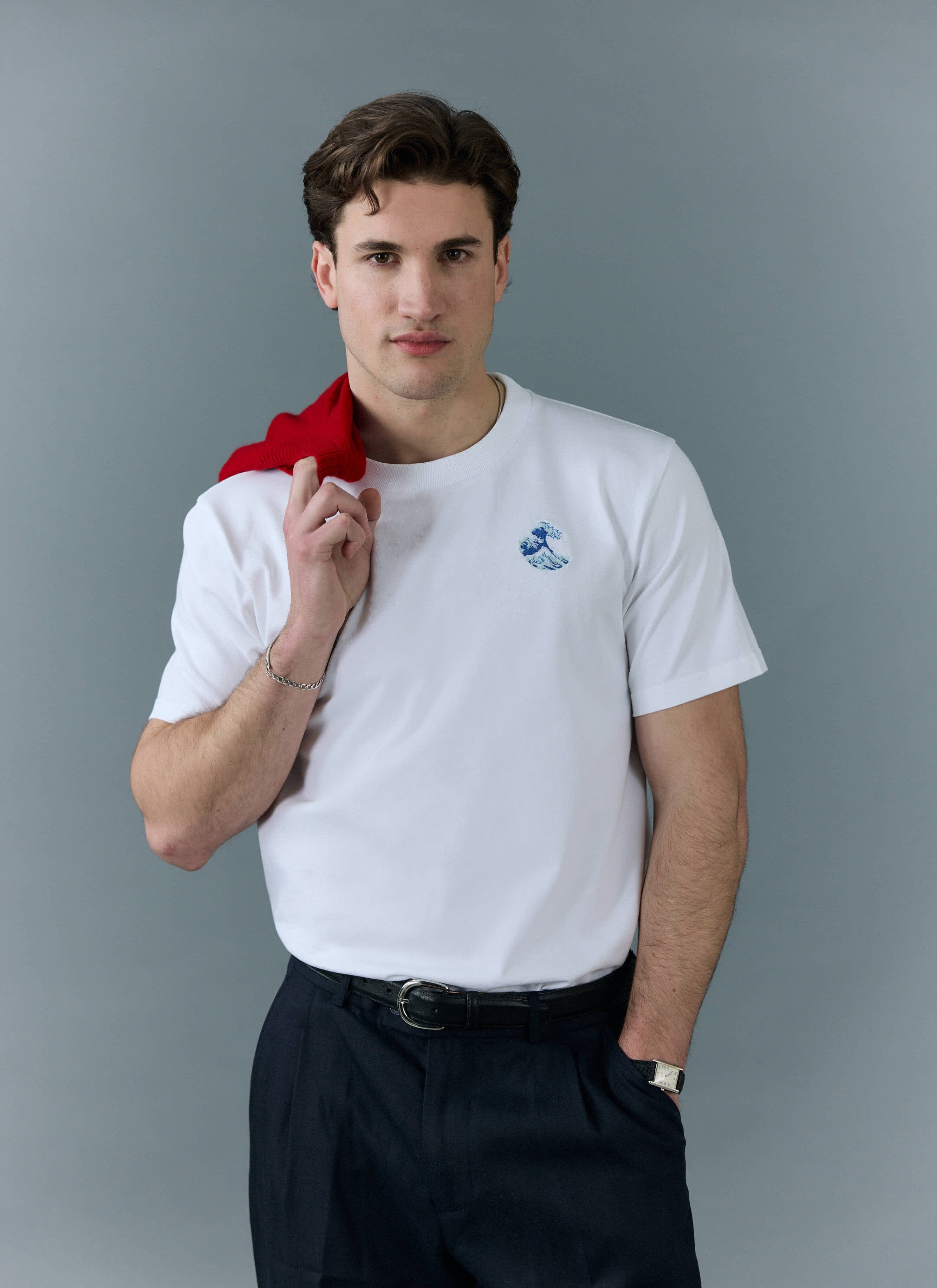 A model wears the white Percival Wave T Shirt with blue embroidery, styled with dark trousers and a red jumper on shoulder.