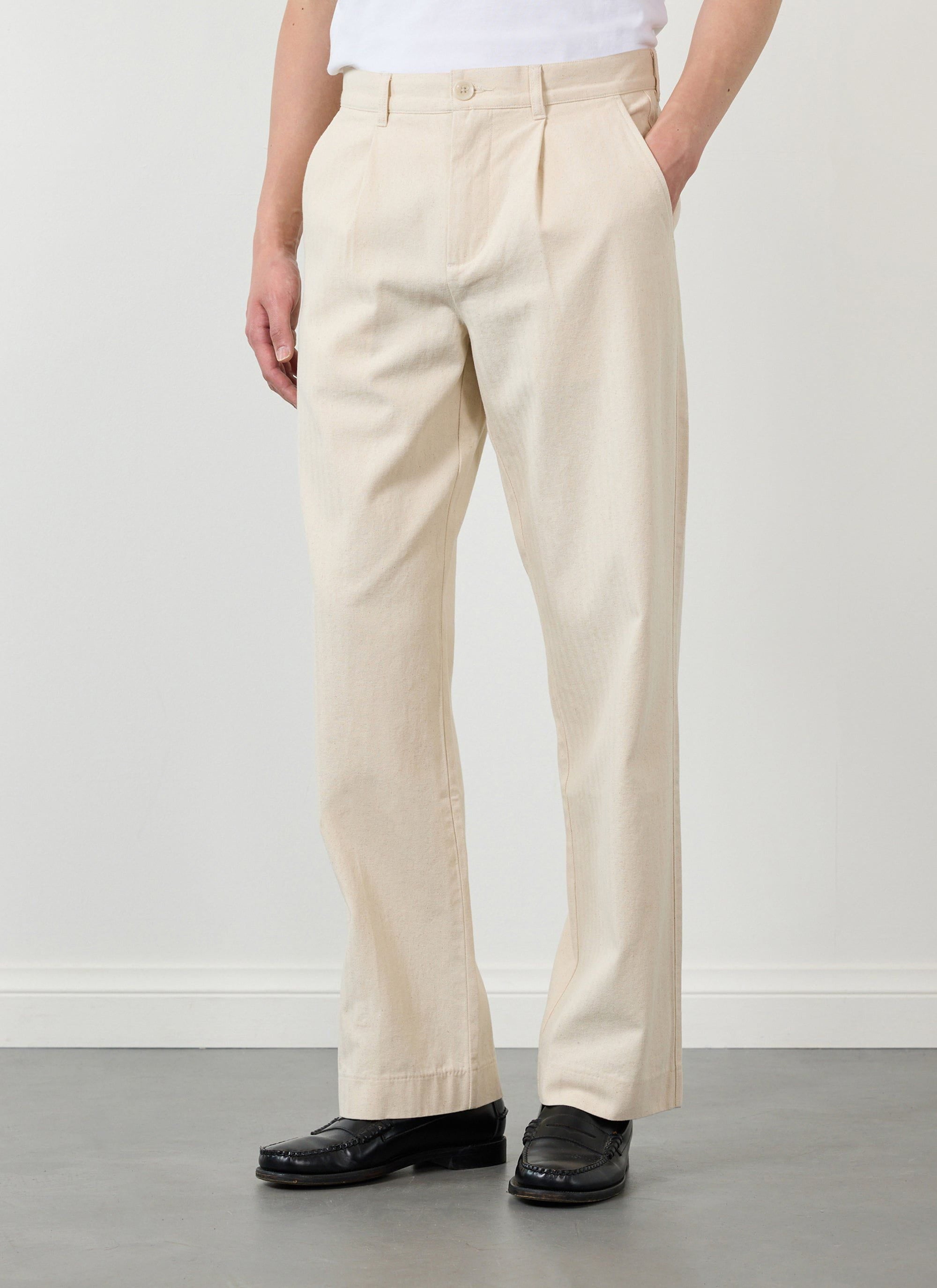 A model wears the Percival Program Pleated Trousers in ecru cotton with black loafers.