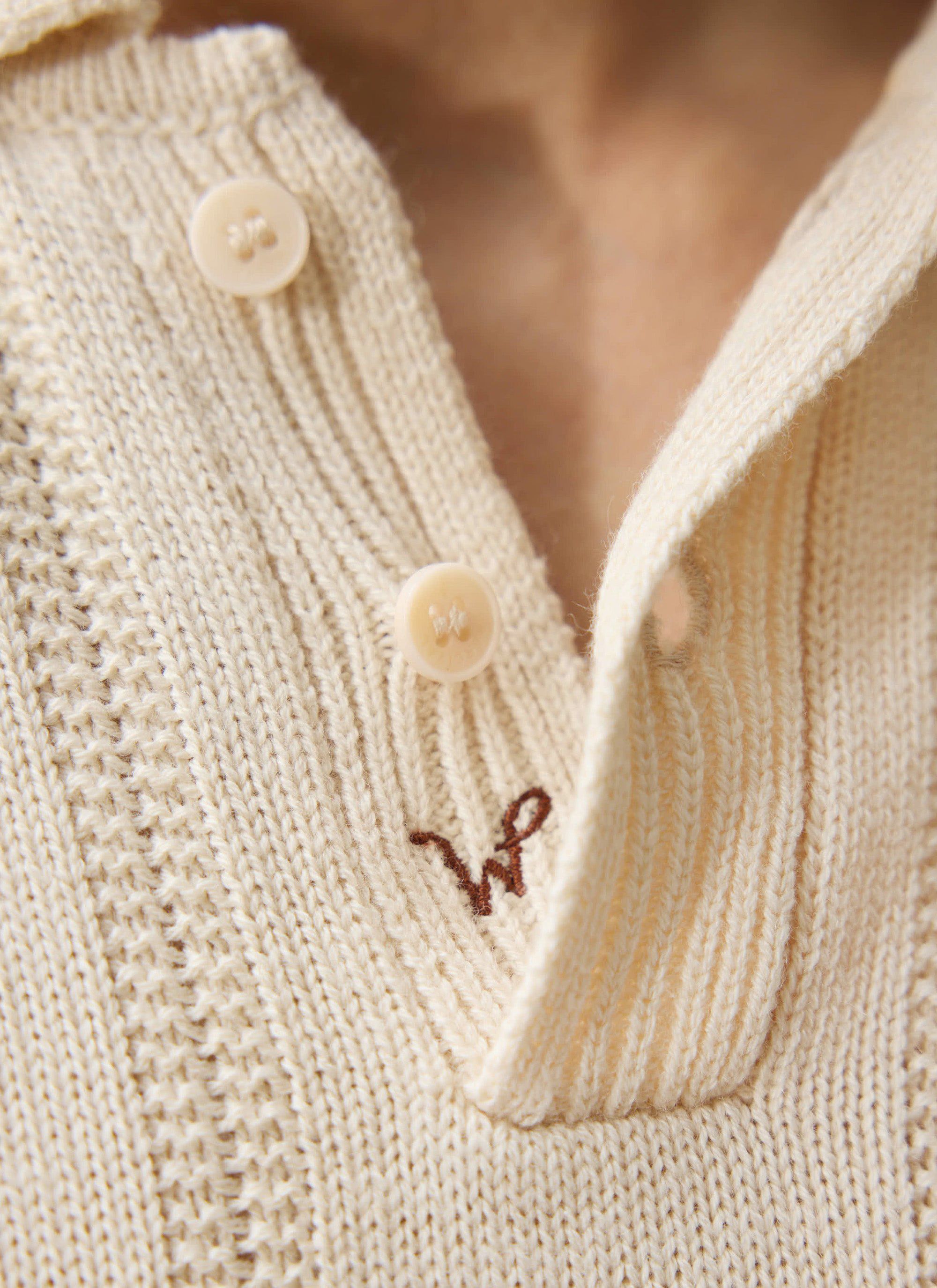 A close-up of the Percival x Warren knitted polo in ecru, showing the button placket and embroidered 'W' logo.