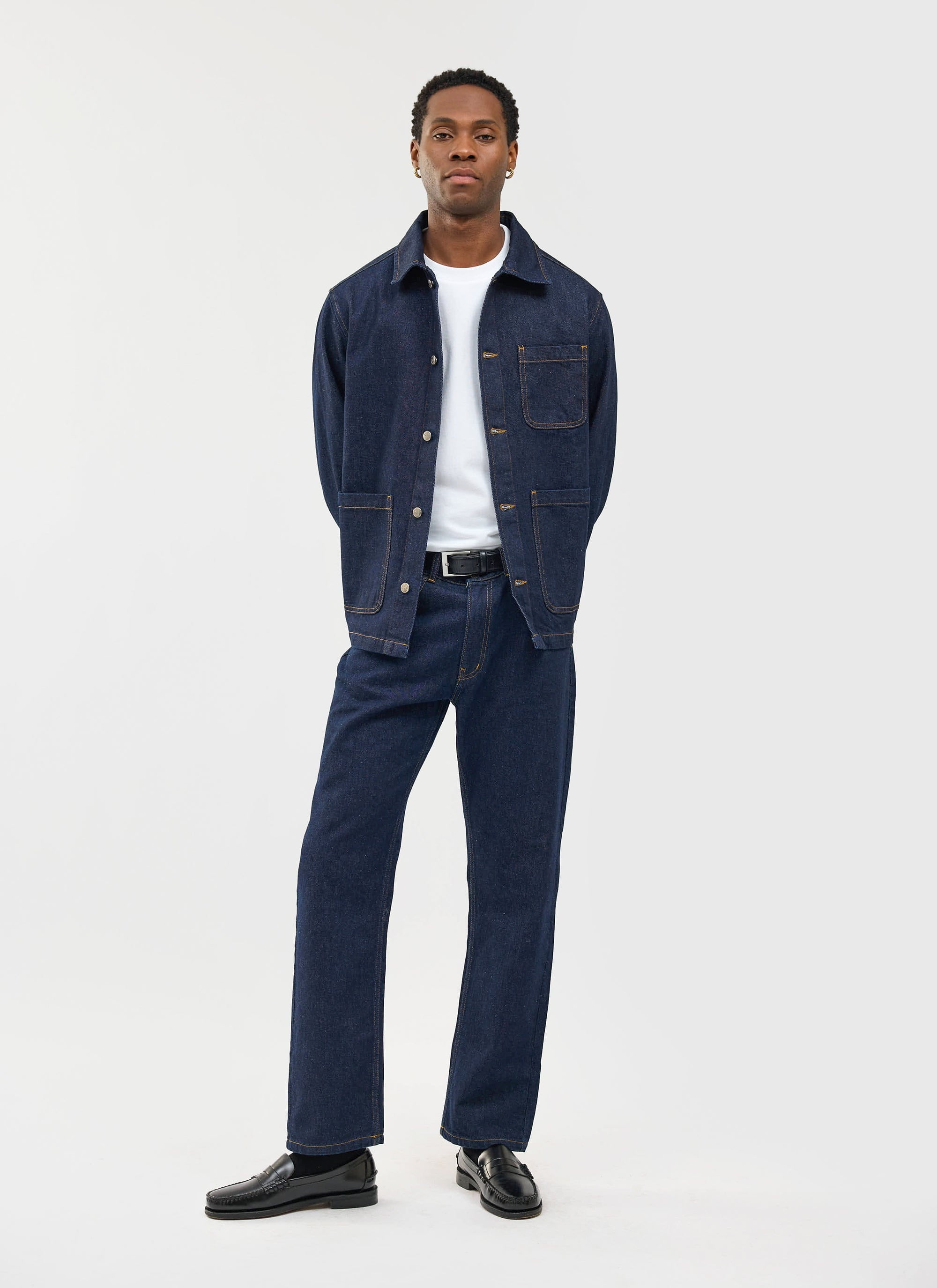 A man models the Percival Gove Chore Jacket in dark indigo denim, paired with matching trousers and a white t-shirt.