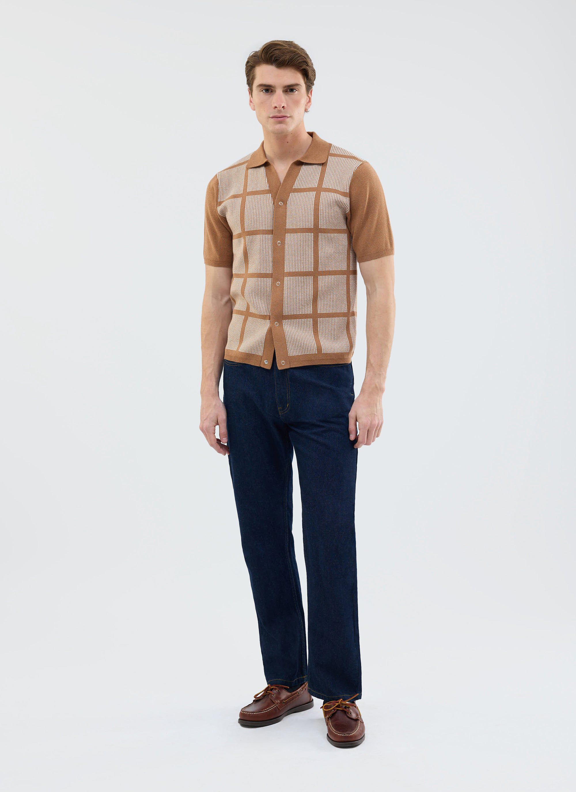 A model wears the Percival Costa Knitted Shirt in tobacco, featuring a square pattern, with dark jeans and boat shoes.