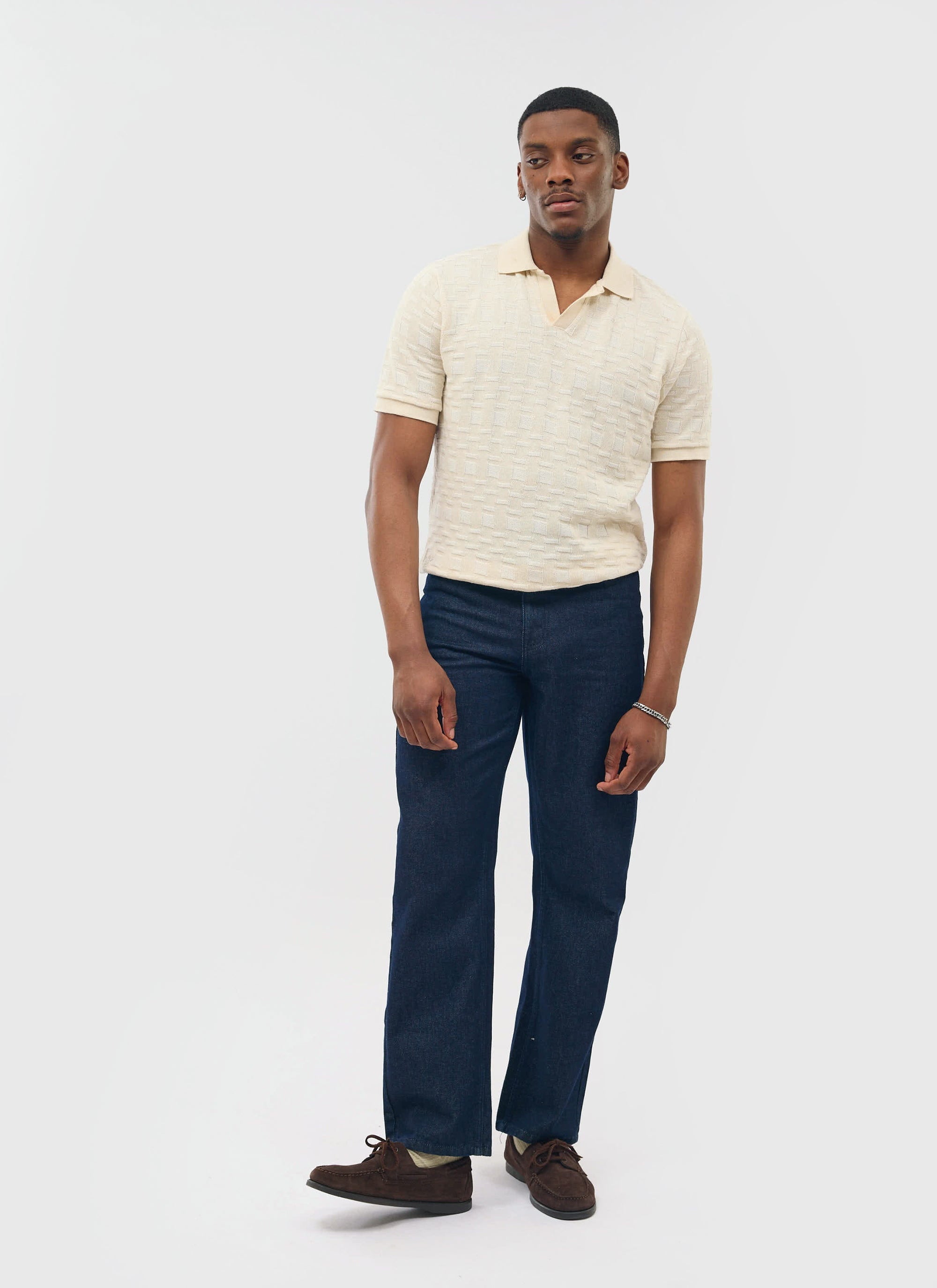 A model wears the Percival Deck Negroni Knitted Shirt in ecru with dark denim jeans and brown suede shoes.