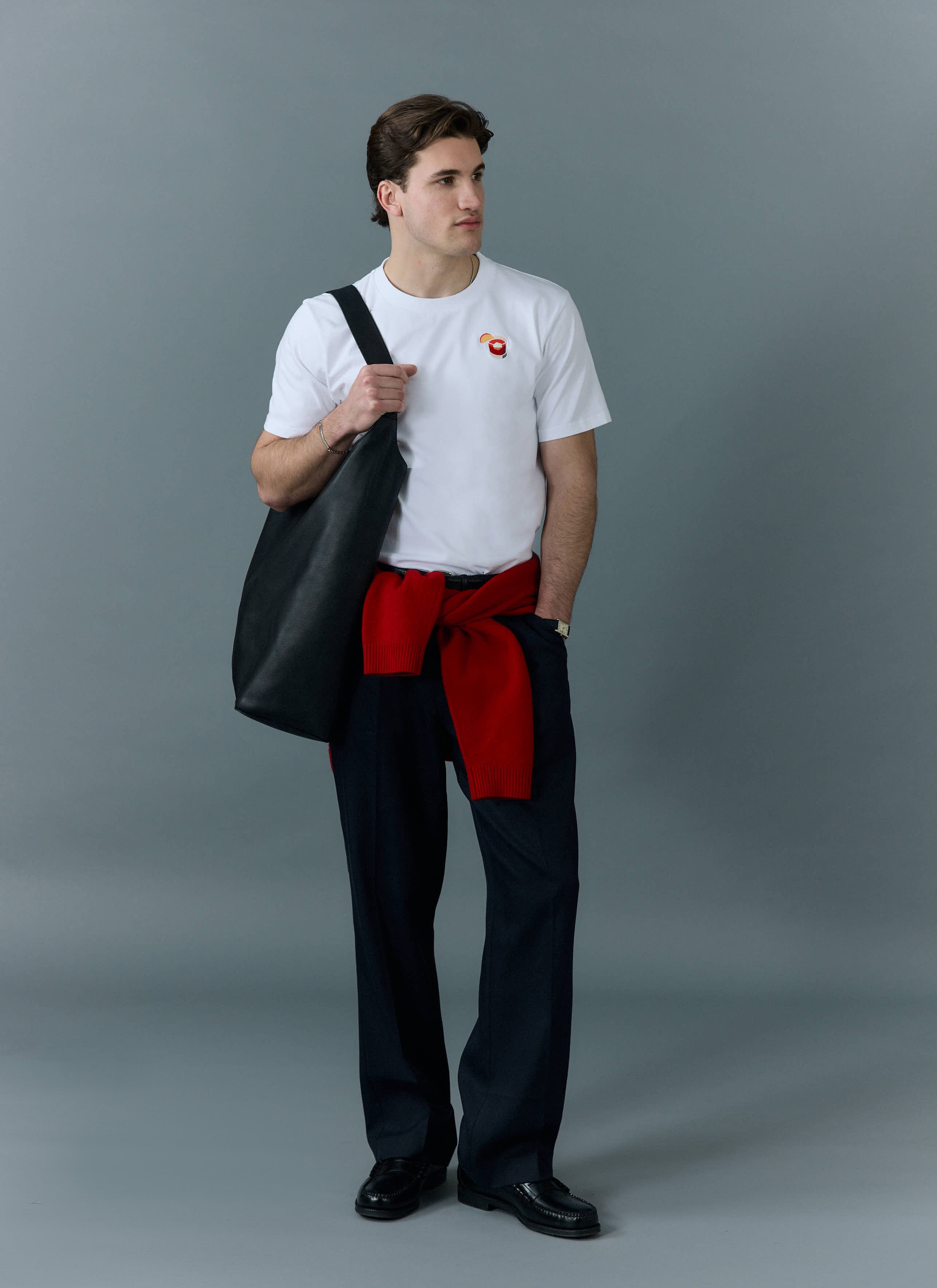 A model wears the white Percival Negroni T-shirt, styled with dark trousers, a red jumper and a black leather tote bag.