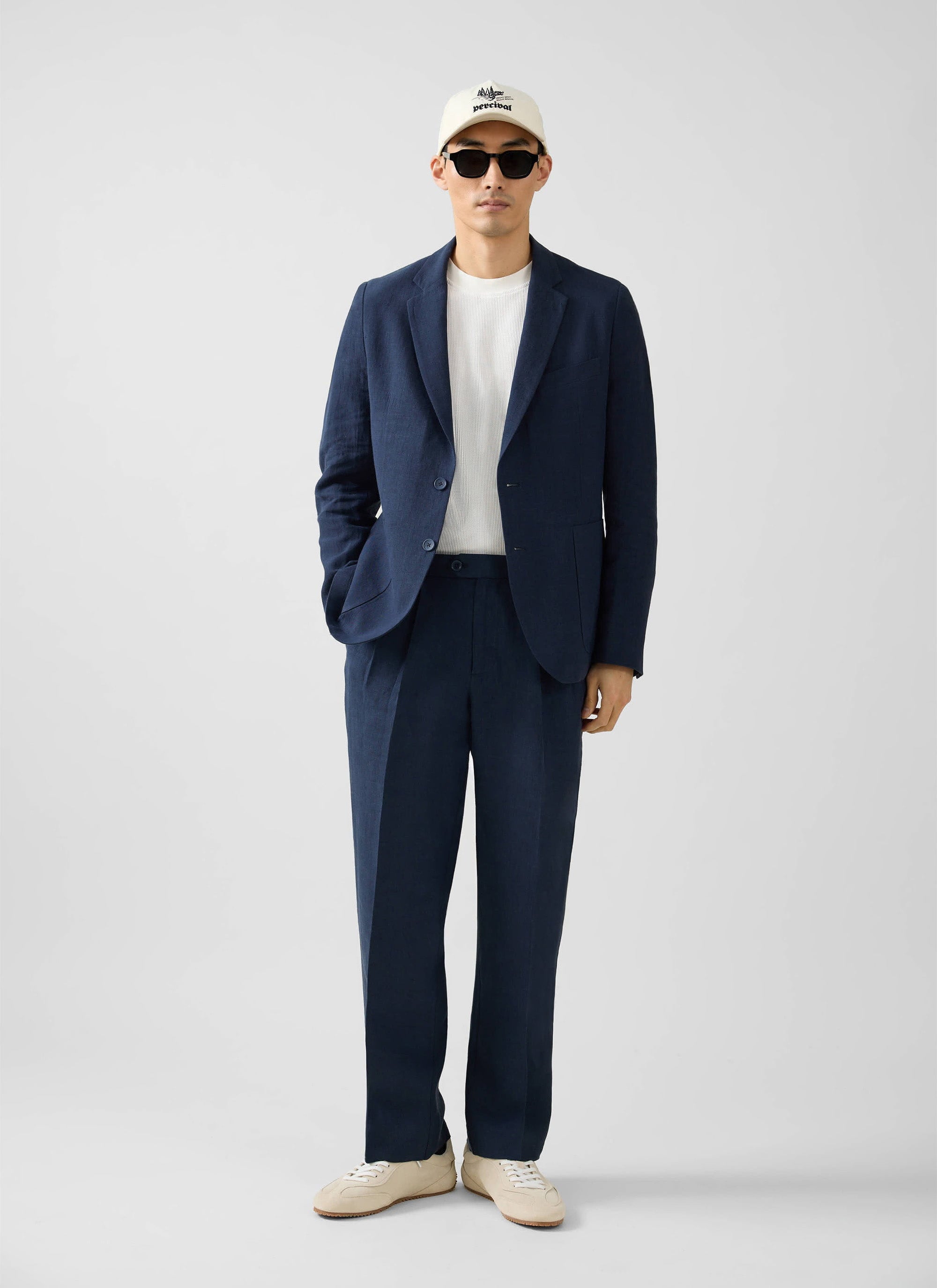 A model wears the Percival navy tailored linen blazer with matching trousers, a white t-shirt, trainers, and a cap.