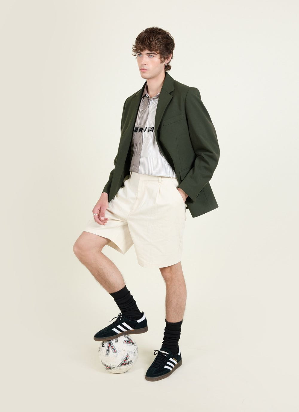 A model wears the Percival forest green tailored blazer with cream shorts and a shirt, resting his foot on a football.