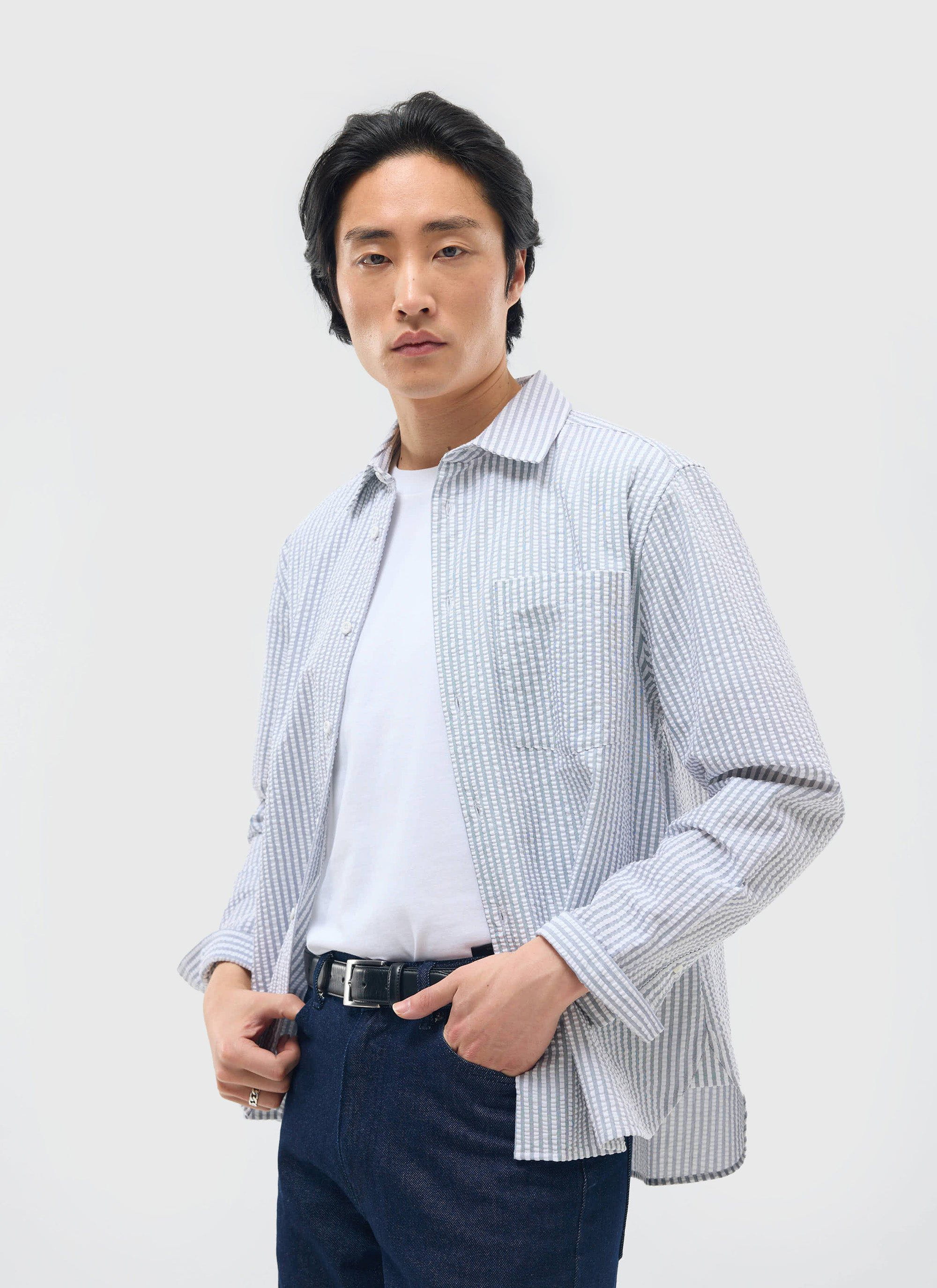 A man models the Percival Seersucker Stripe Pearce Shirt in grey, worn open over a white t-shirt and dark blue jeans.