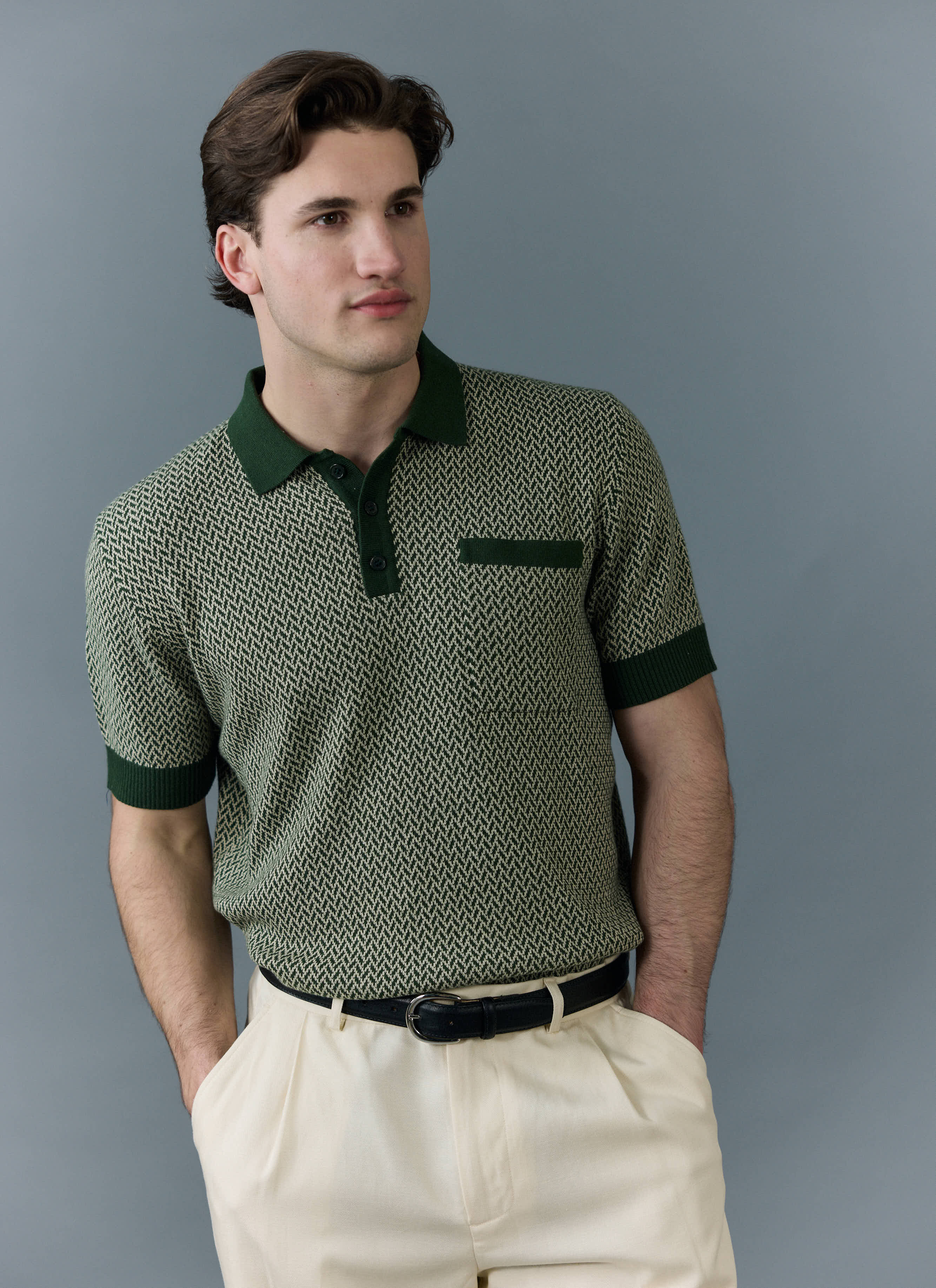 Model wears the Percival Casa Martini knitted polo in forest green, paired with cream trousers against a grey background.