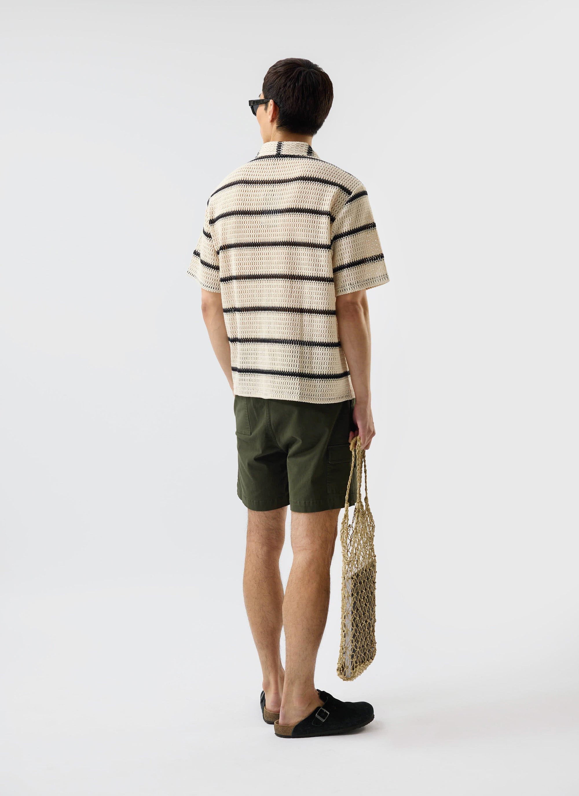 A model, seen from behind, wears the Percival Crochet Groundsman Cuban Shirt in ecru with black stripes and green shorts.