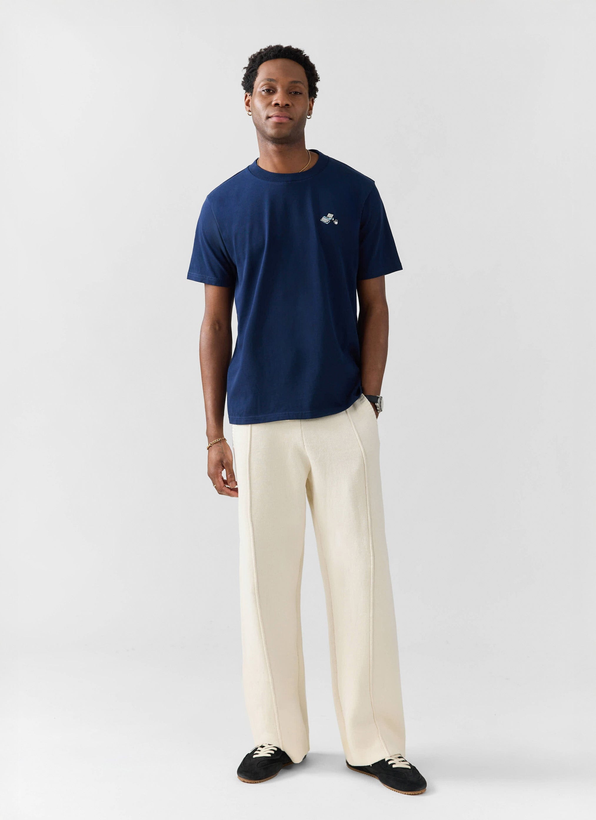 A model wears the navy Typewriter Embroidered T-shirt from Percival, styled with wide-leg cream trousers and black trainers.