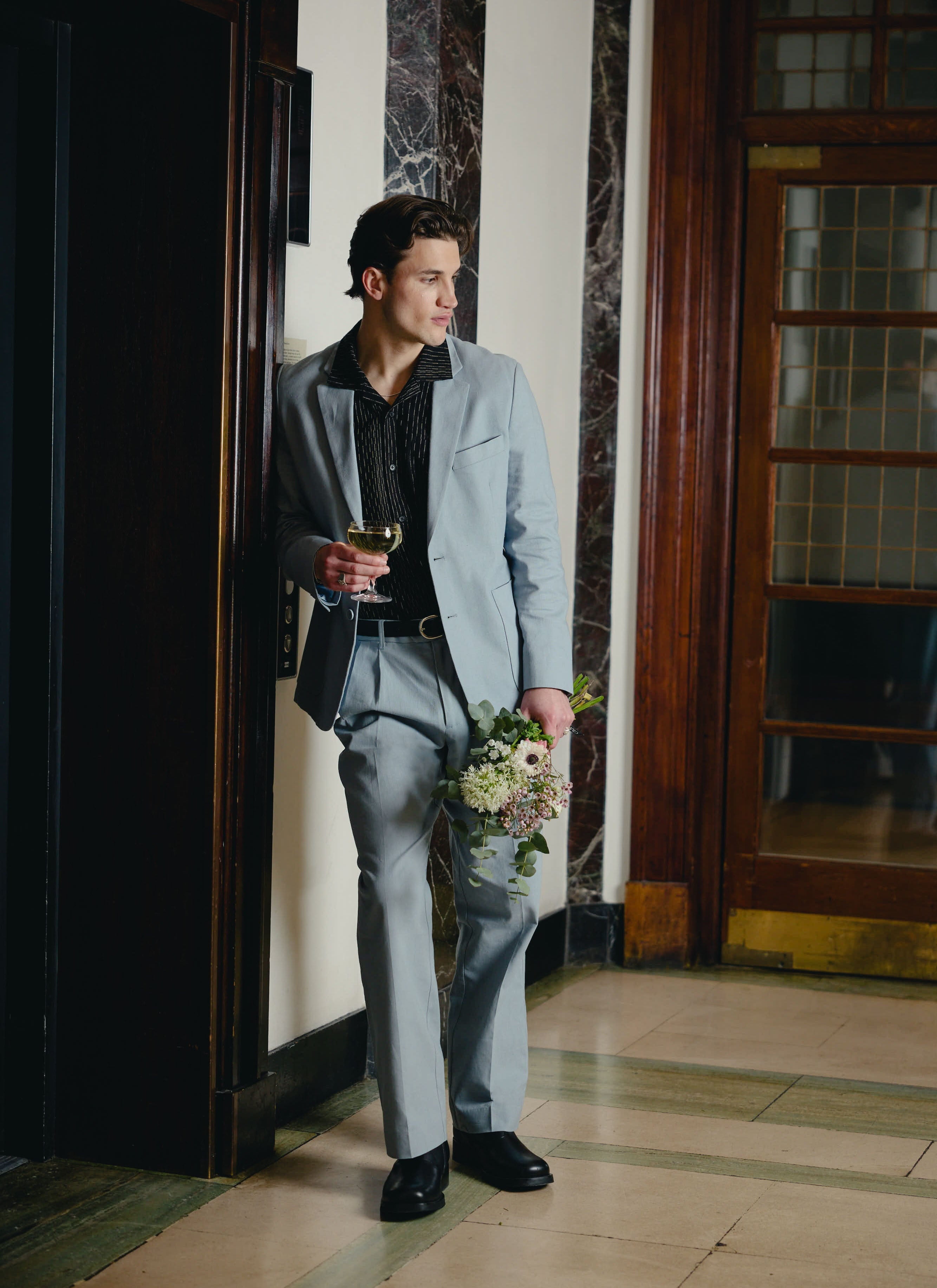 A model wears the Percival Tailored Linen Blazer and matching trousers in light blue, holding flowers and a drink.
