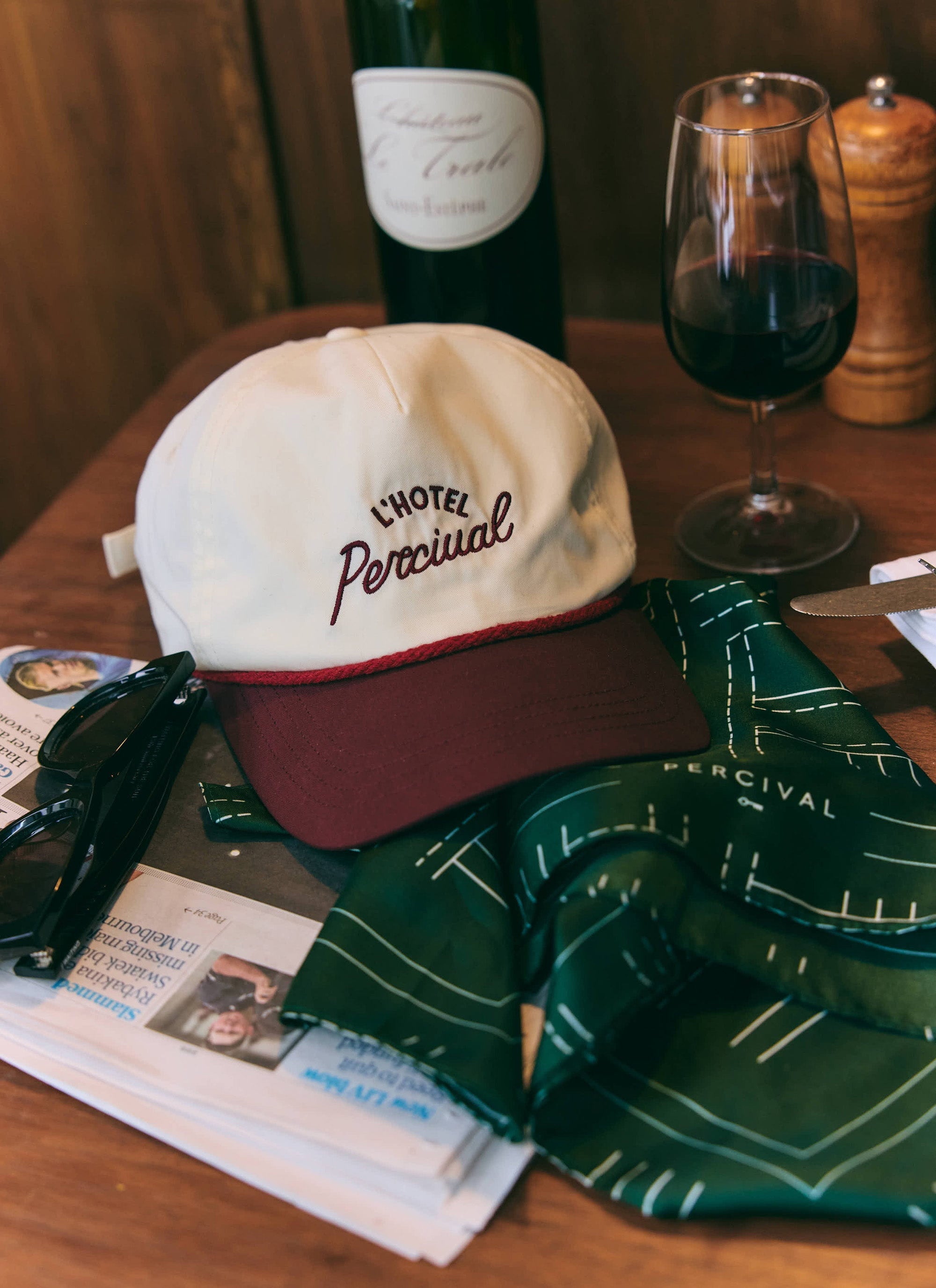 The ecru and burgundy L'Hotel Percival soft trucker cap rests on a table, styled with a glass of red wine and a newspaper.