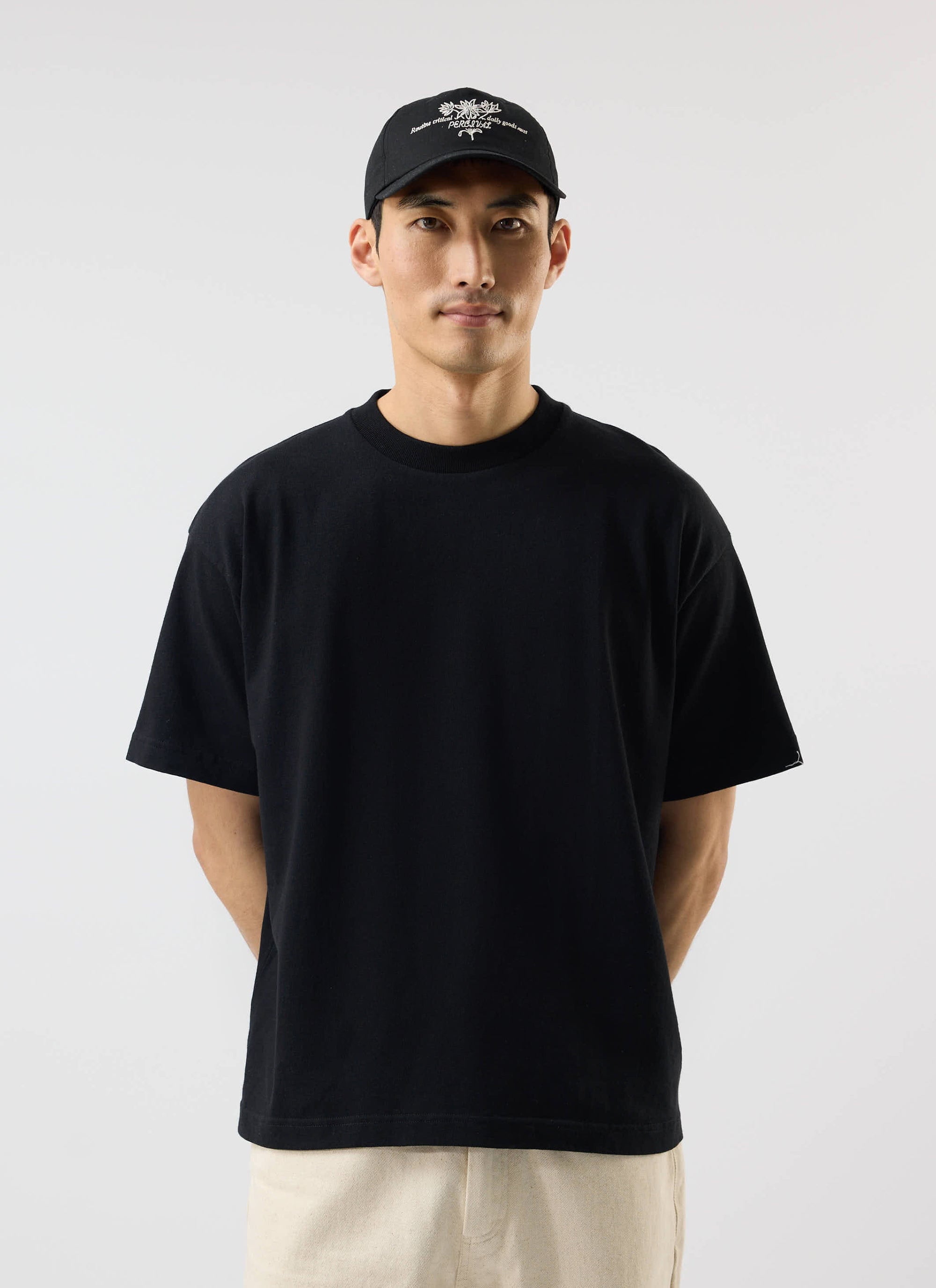 A model wears the Percival oversized t-shirt in black, made from organic cotton, with a black cap and cream trousers.