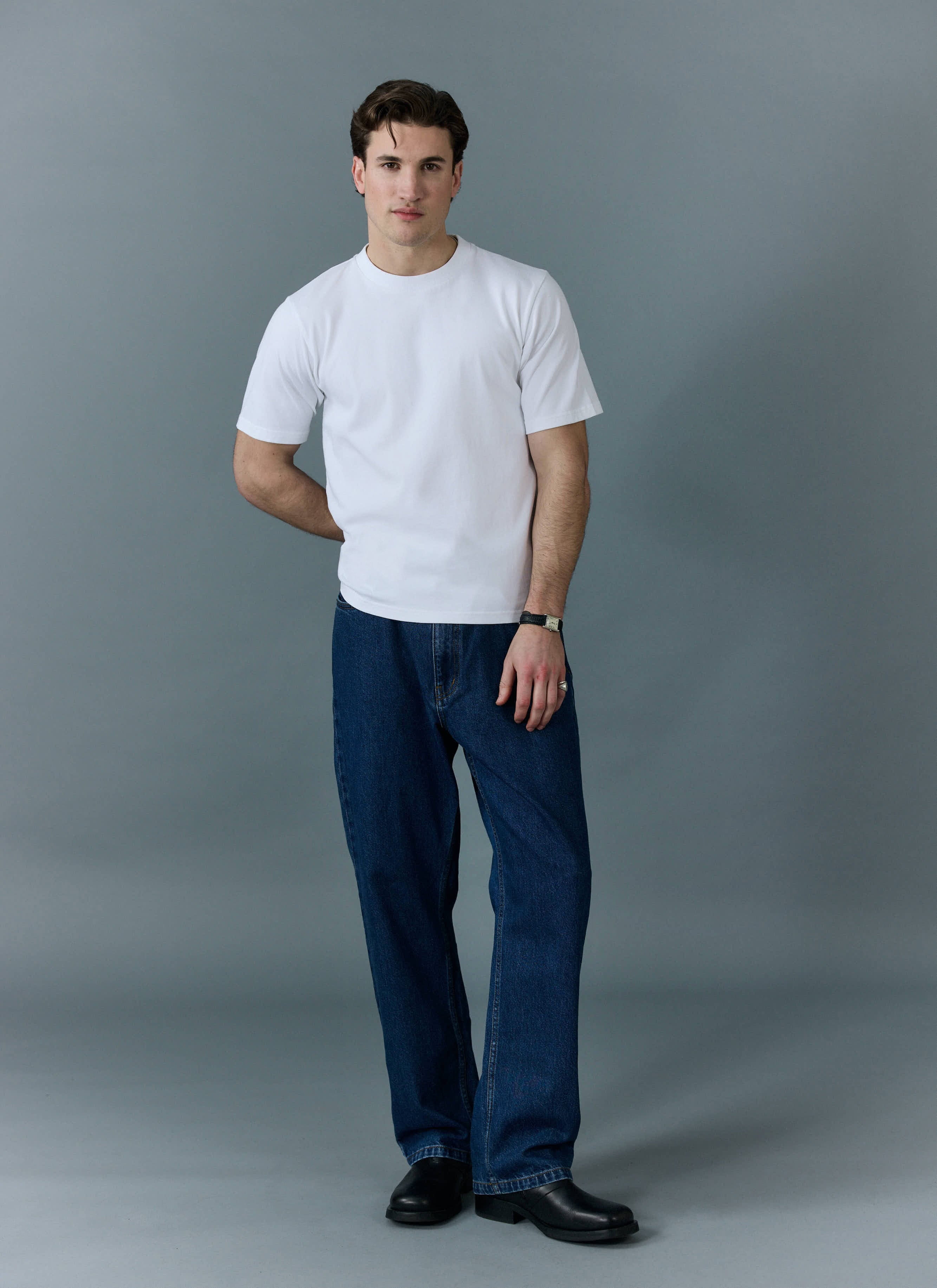 A model wears the Percival white Cropped Regular Fit T-Shirt with dark blue jeans and black shoes, against a grey background.