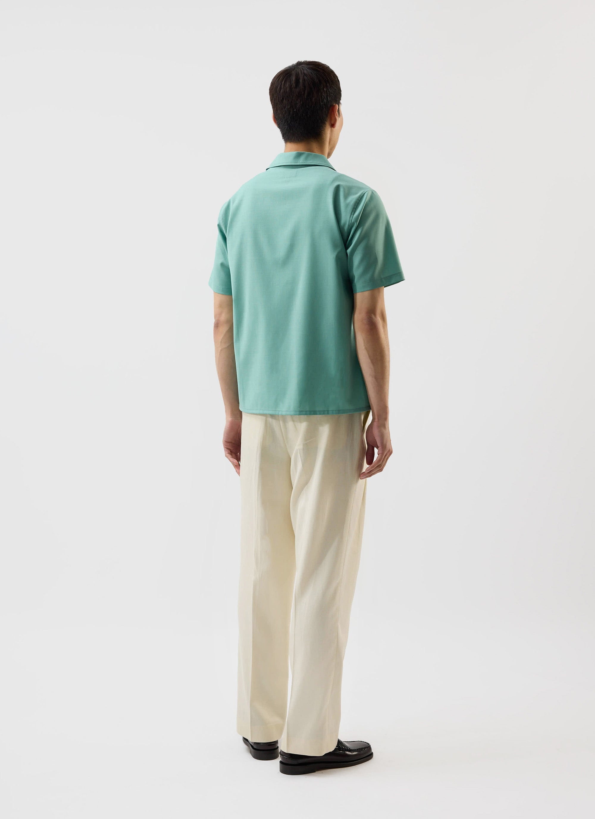 A model, seen from behind, wears the mint green Percival x Warren Drape Cuban Shirt with cream trousers.