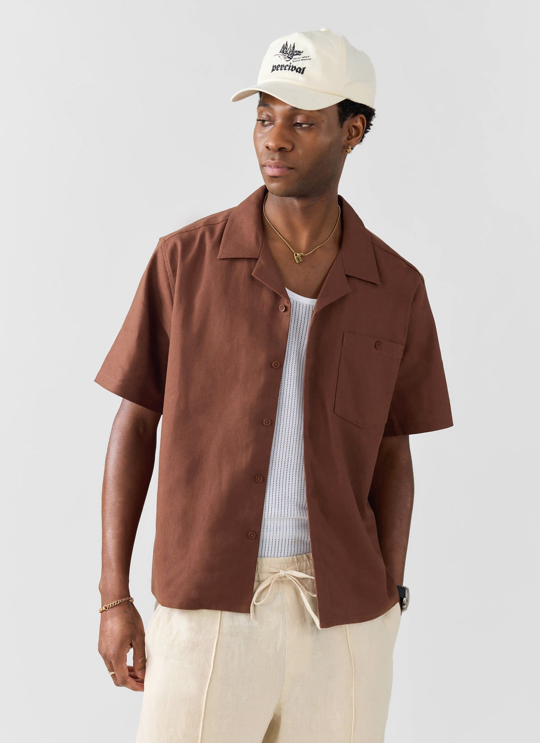 A model wears the Percival brown linen Cuban shirt, open over a white vest, with cream trousers and a Percival baseball cap.