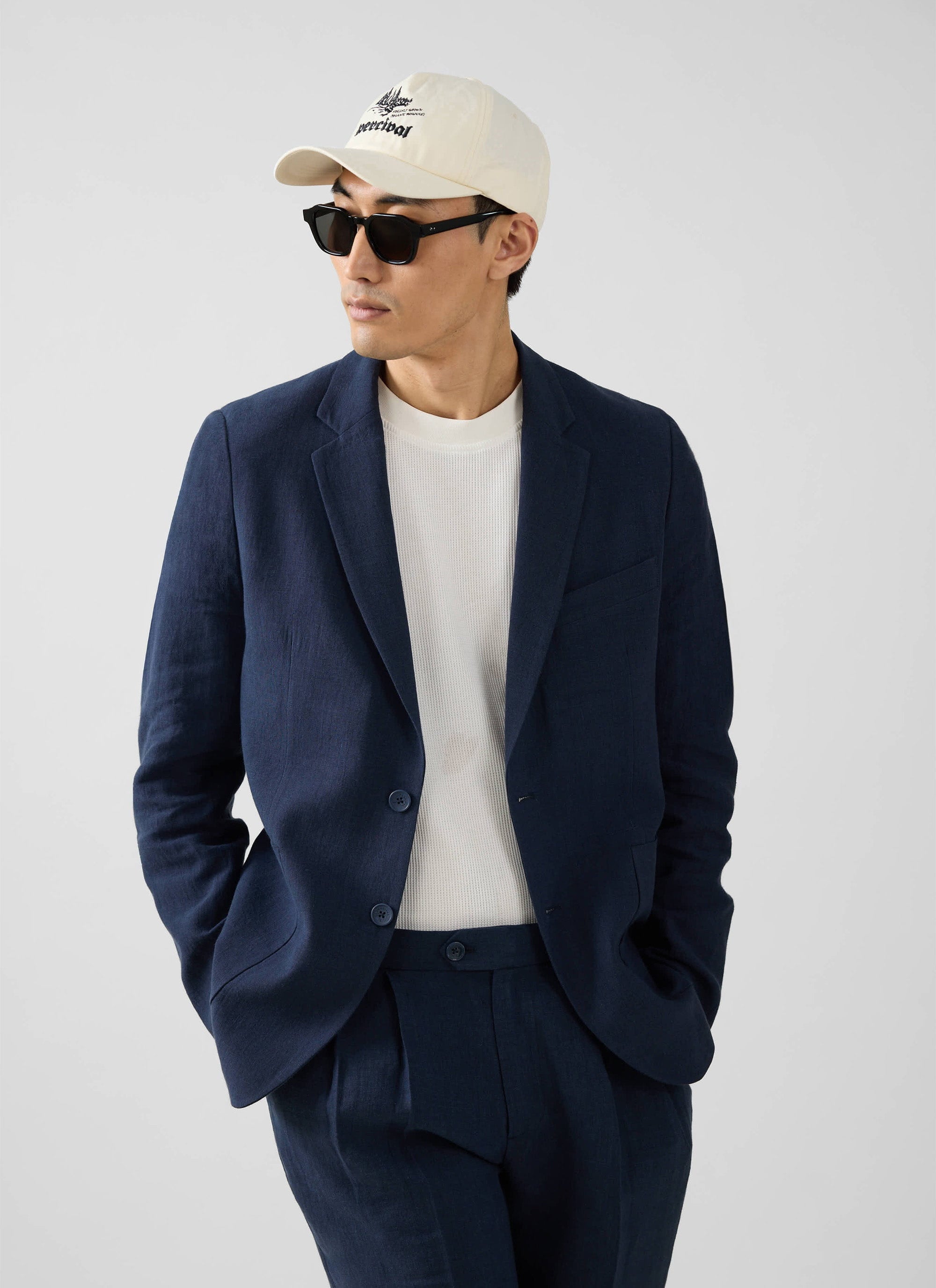Model wears the Percival navy linen blazer with matching trousers, a white t-shirt, sunglasses and a Percival cap.
