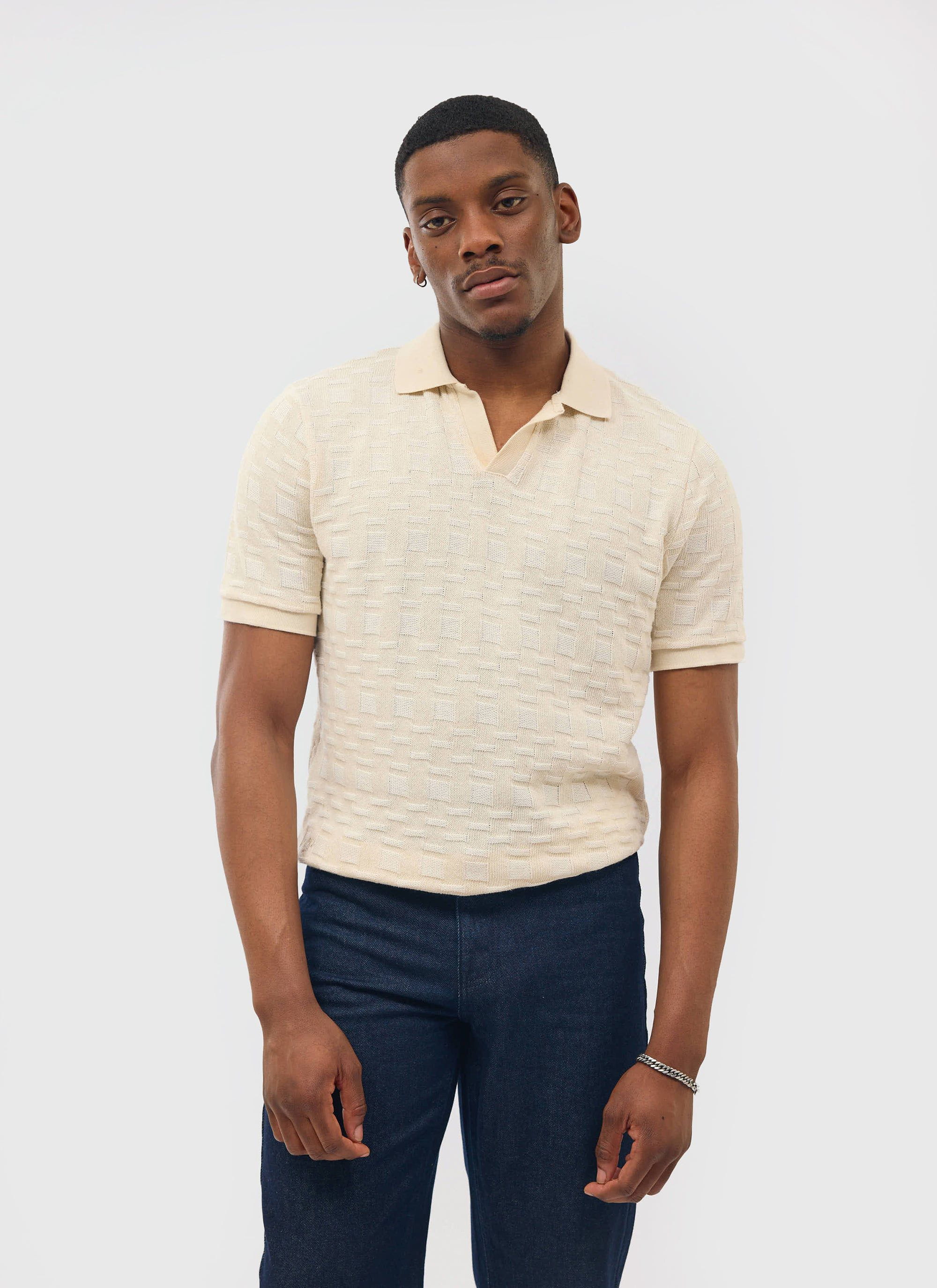 A man models the Percival Deck Negroni knitted polo shirt in ecru, paired with dark blue jeans in a studio setting.