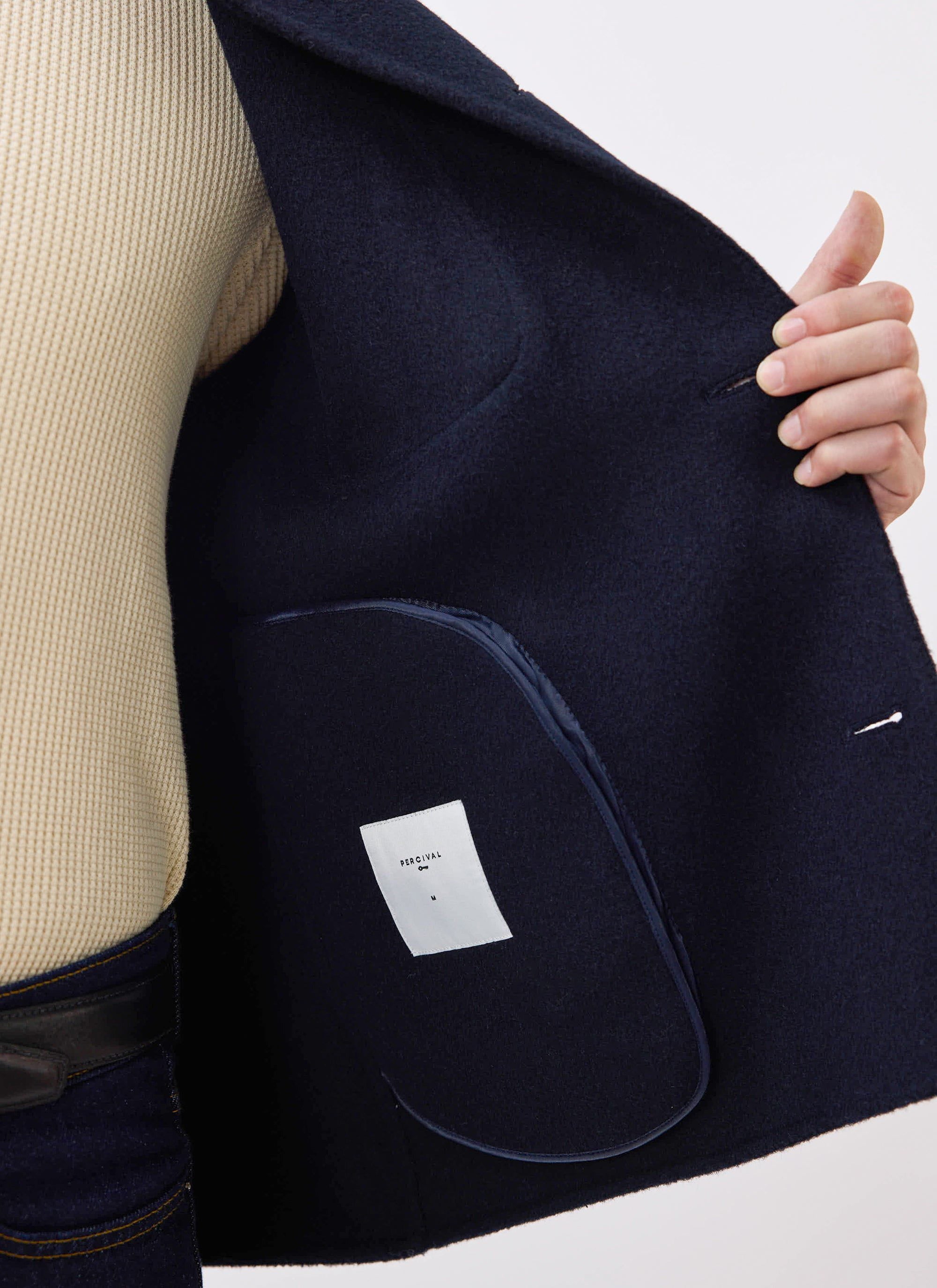 A model shows the internal pocket and branding on the navy Heavy Wool Overshirt from Percival, layered over a cream knit.