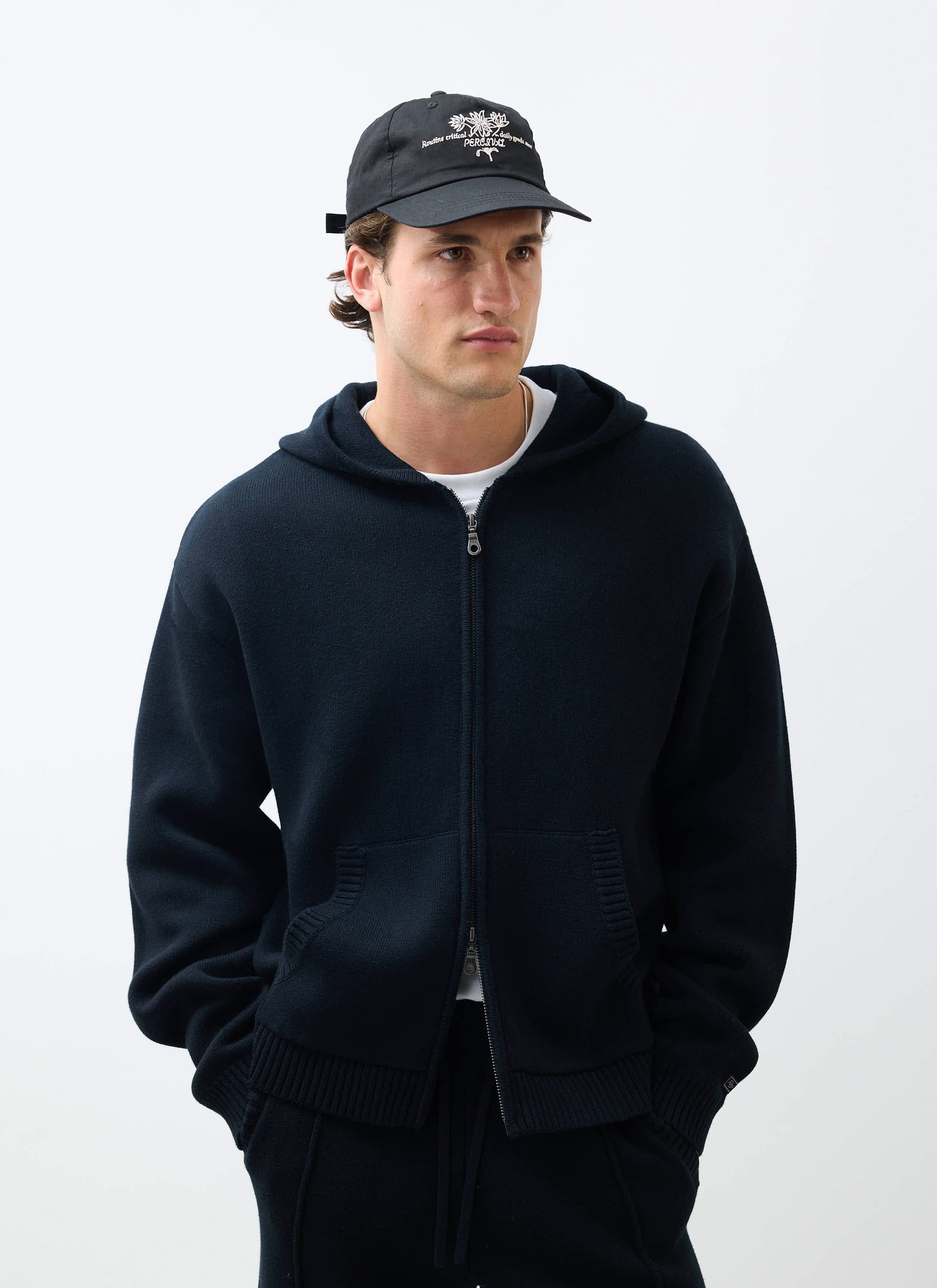 A man models the black Percival Chronicle Knitted Hoodie, a cotton zip-up style, worn with a cap in a studio.