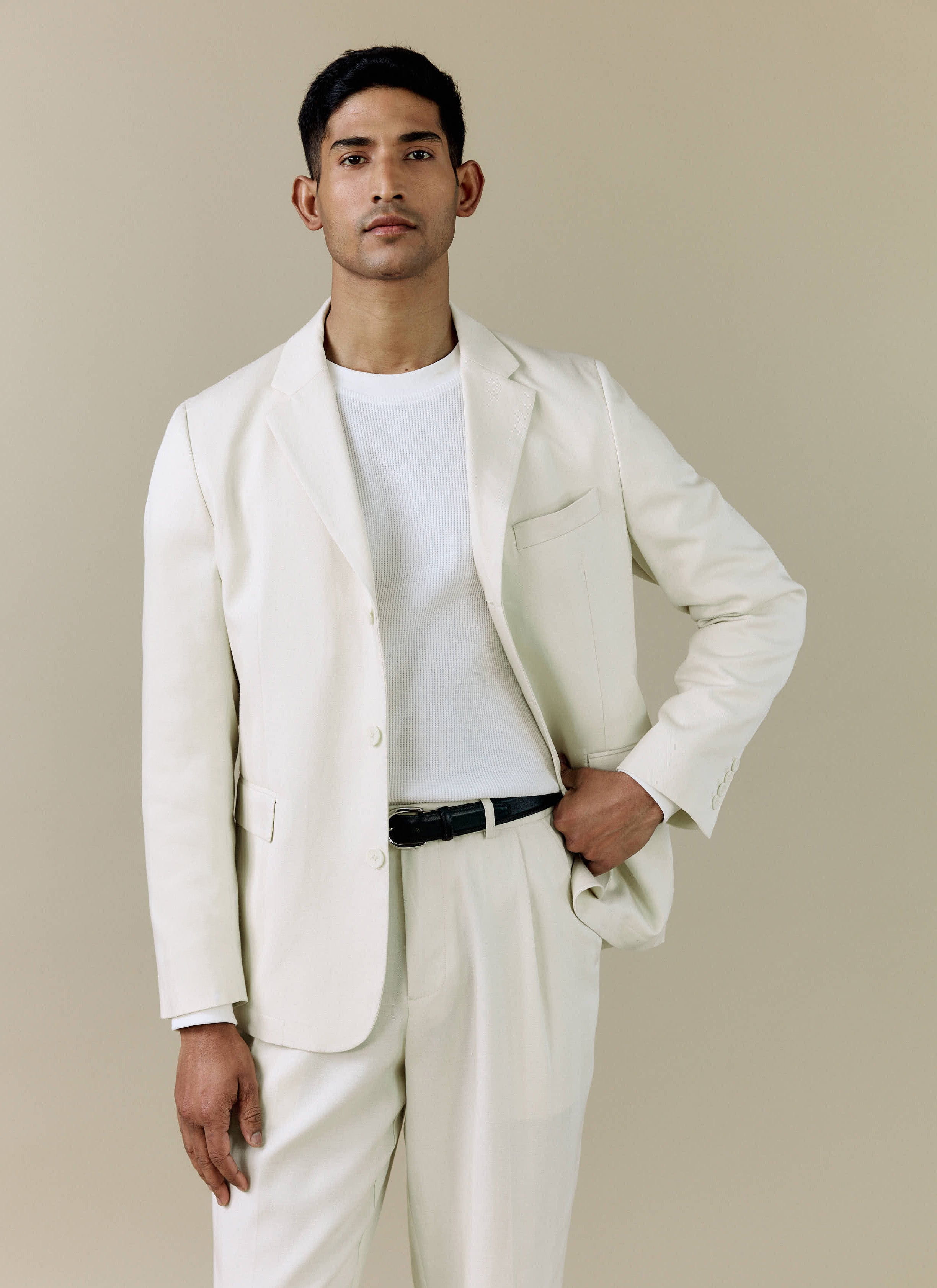 A model wears the Percival Pleated Tailored Trousers in cream, styled with the matching blazer and a white ribbed t-shirt.