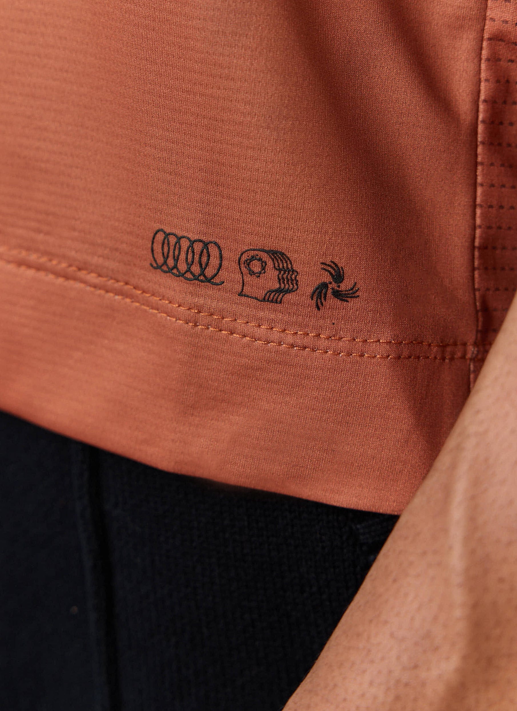 Close-up detail of the hem and black graphic logos on the rust-coloured PCVL Performance Top from Percival.