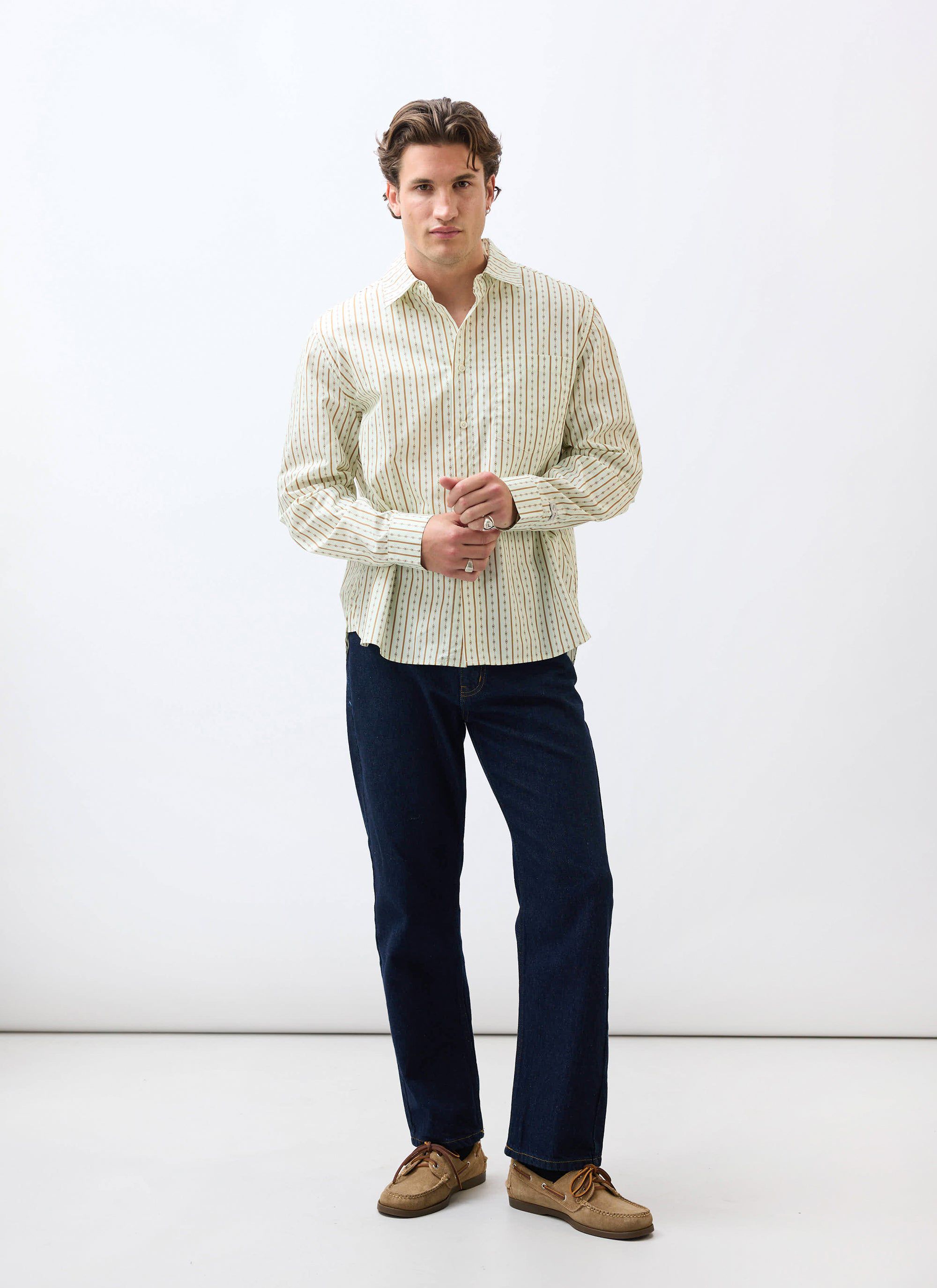 A model stands against a white background wearing the Percival Binding Oversized Pearce Shirt in ecru with dark jeans.