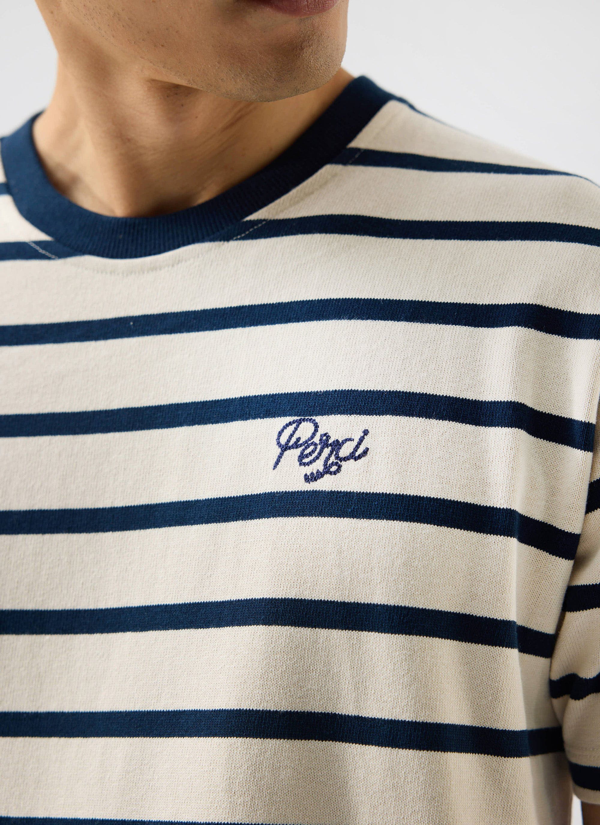 Close up of a model wearing the Percival Breton Stripe T Shirt in ecru and navy, showing the chest embroidery.