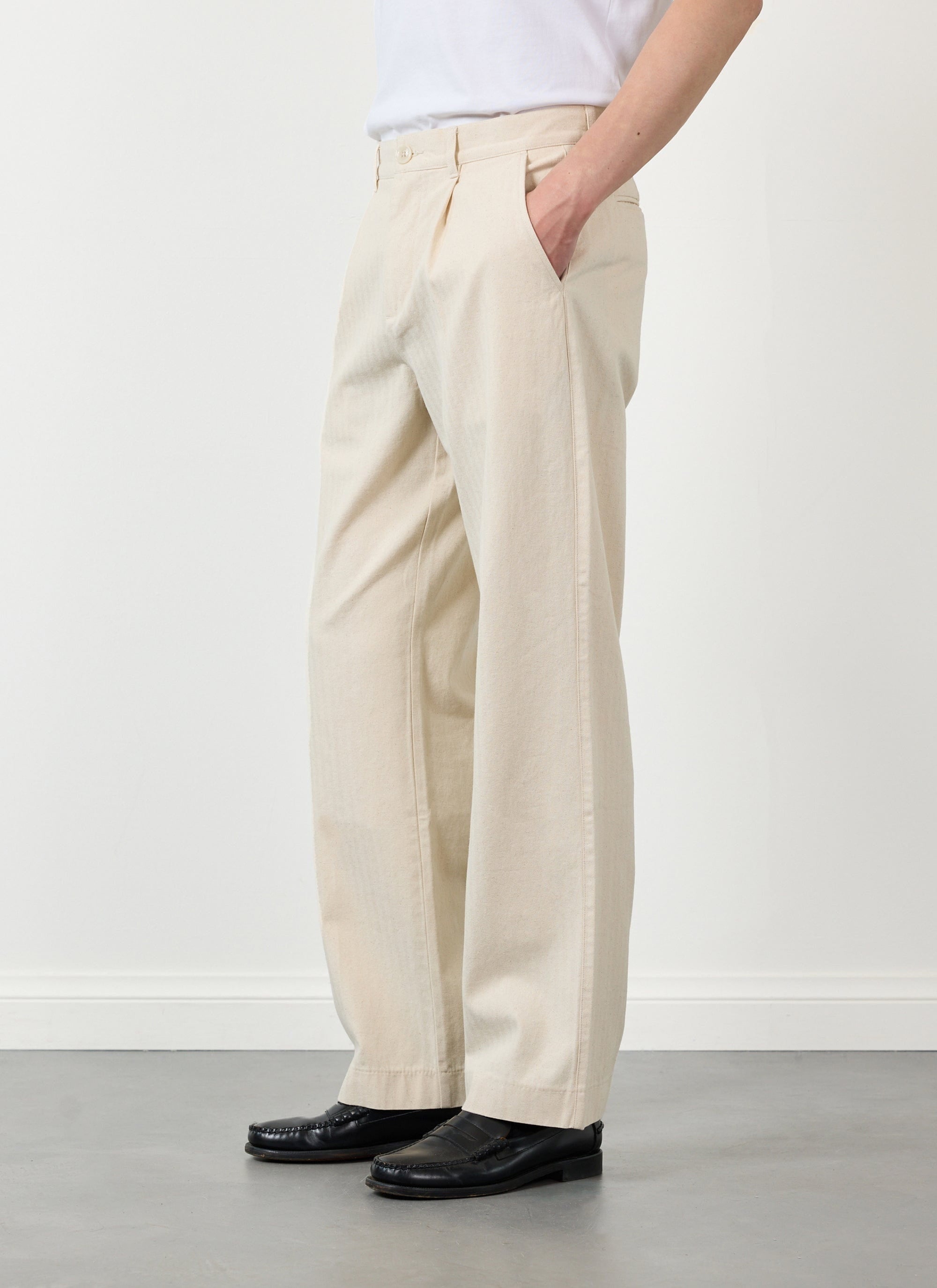 Model from the side wearing Percival ecru pleated cotton trousers, a white tee and black loafers with hand in pocket.
