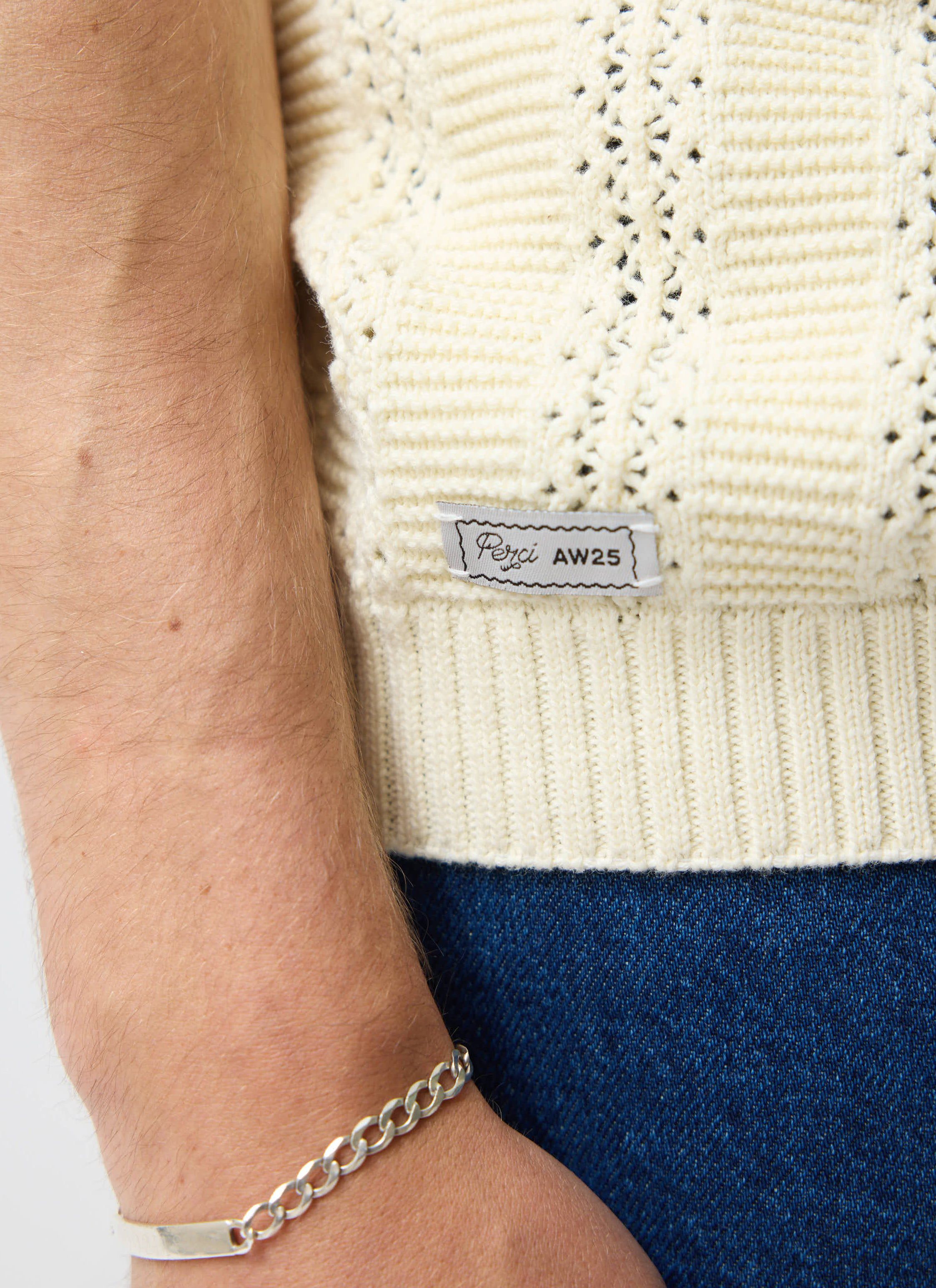 A close up of the Percival Addendum Knitted Polo Shirt in ecru cream, showing the tonal knit detail and Perci AW25 side tab.